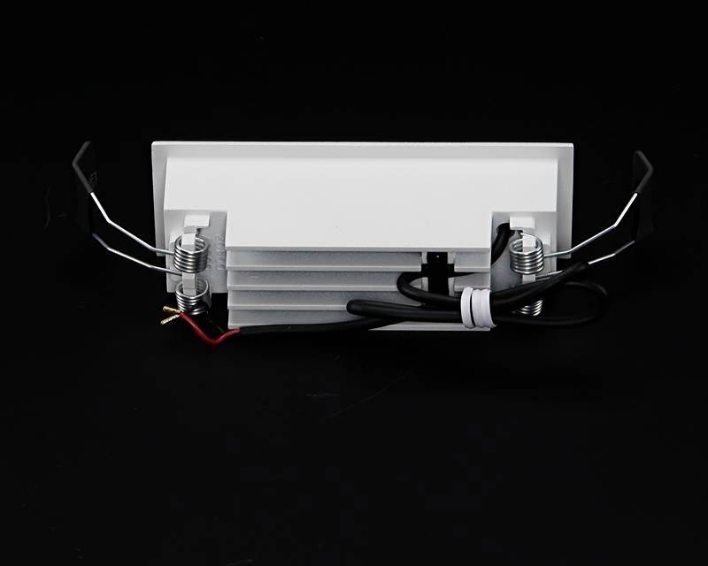 Obdĺžniková biela stropná lampa s vyčnievajúcimi kovovými sponkami po stranách a čiernym napájacím káblom na čiernom pozadí.