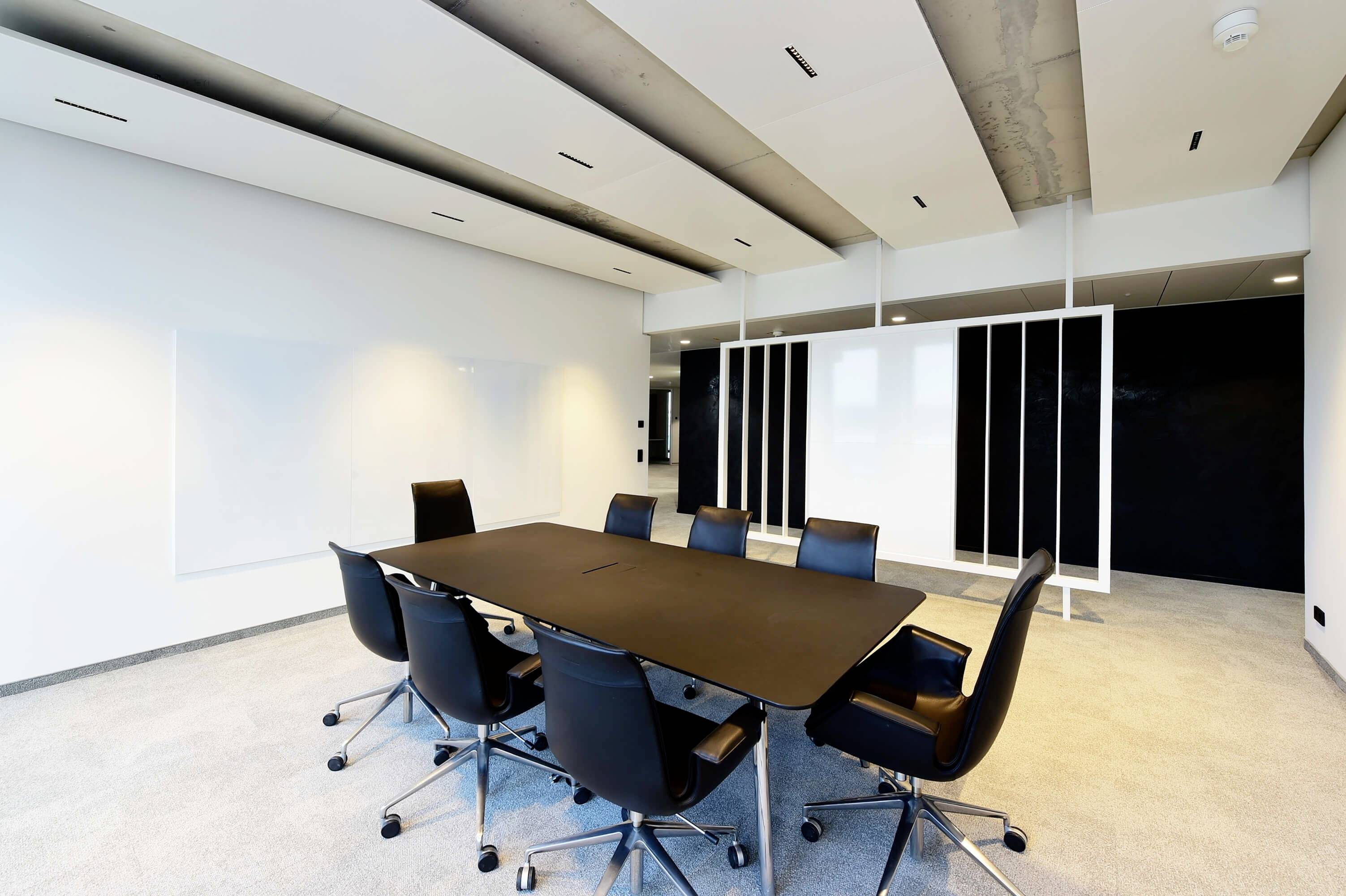 Moderná, prázdna konferenčná miestnosť s veľkým čiernym stolom a ôsmimi čiernymi stoličkami na svetlom koberci.