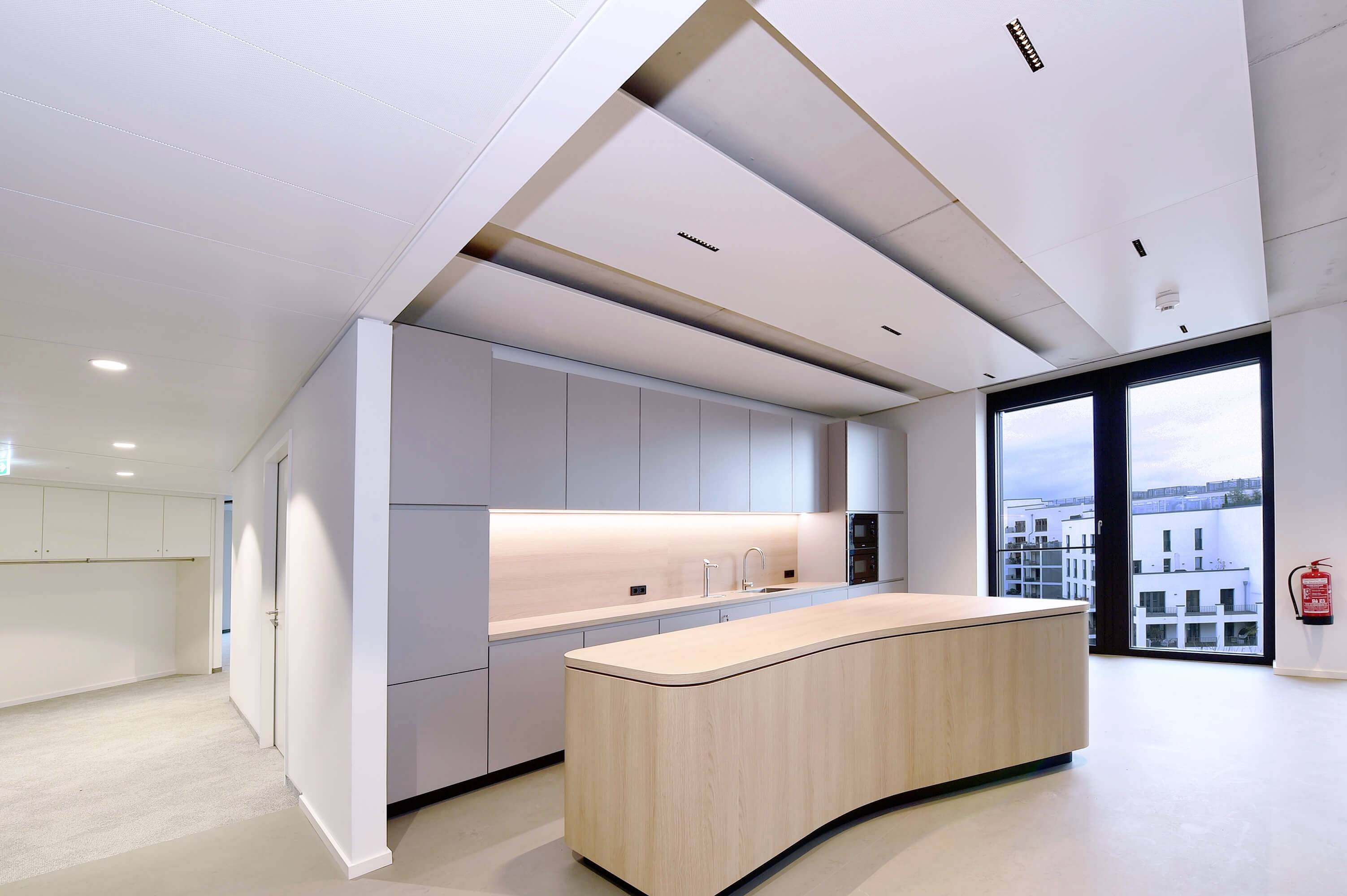 Moderná kuchyňa s oblúkovou drevenou ostrovčekovou stenou, svetlými vstavaným skrinkami a veľkou presklenou stenou s výhľadom na budovu v pozadí.