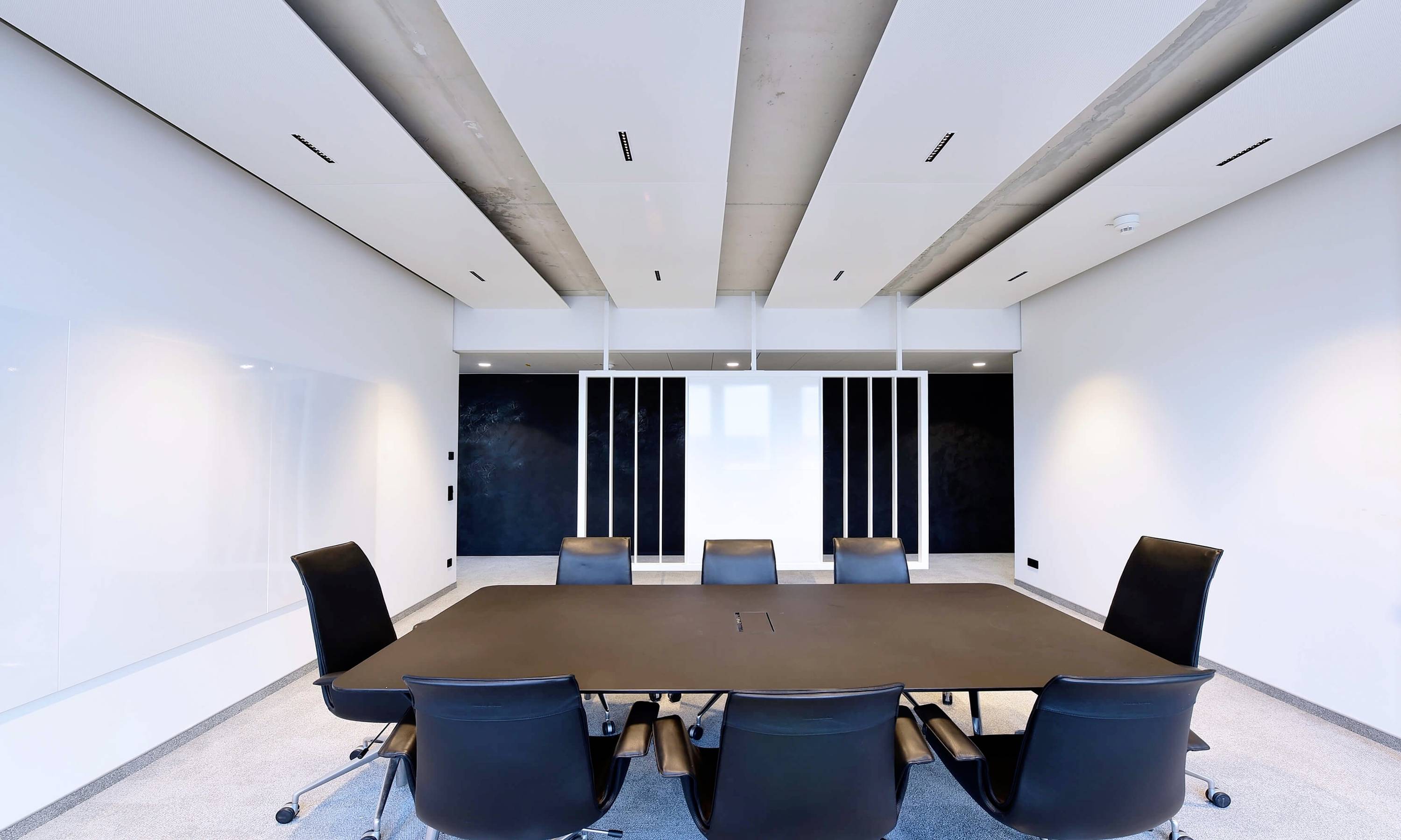 Konferenčná miestnosť s veľkým stolom a siedmimi stoličkami. Steny sú biele, s čiernymi tabuľami na čelnej strane. Priestor pôsobí moderne a minimalisticky.