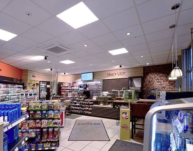 Moderný snack bar s policami plnými sladkostí a nápojov. Zákazník stojí pri pokladni, v pozadí je tabuľa s nápisom 'Snack Shop'.