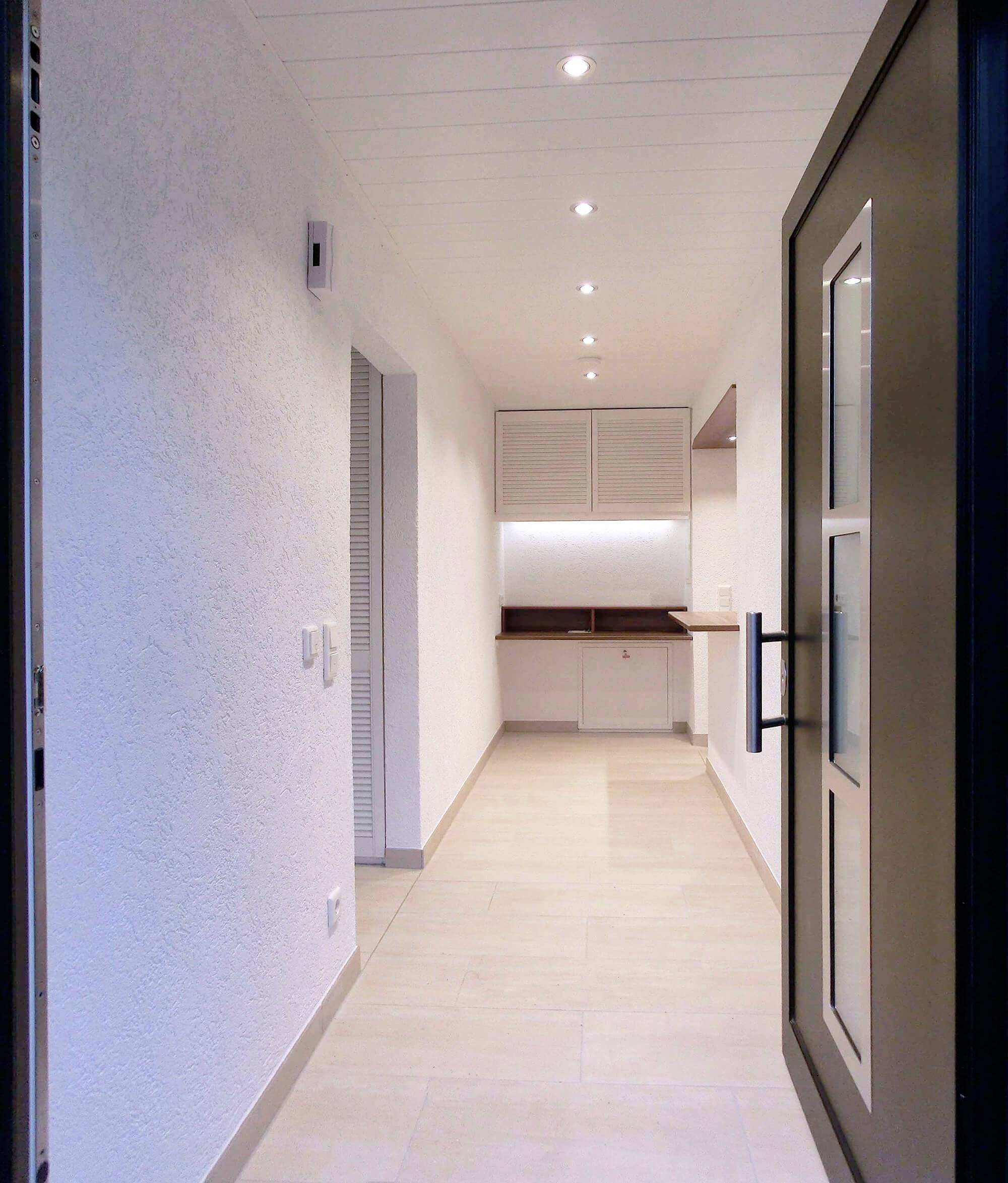 Otvorené dvere vedú do úzkeho, moderného chodníka so svetlou podlahou a bielymi stenami. Na konci chodníka je vidieť kuchynskú linku.