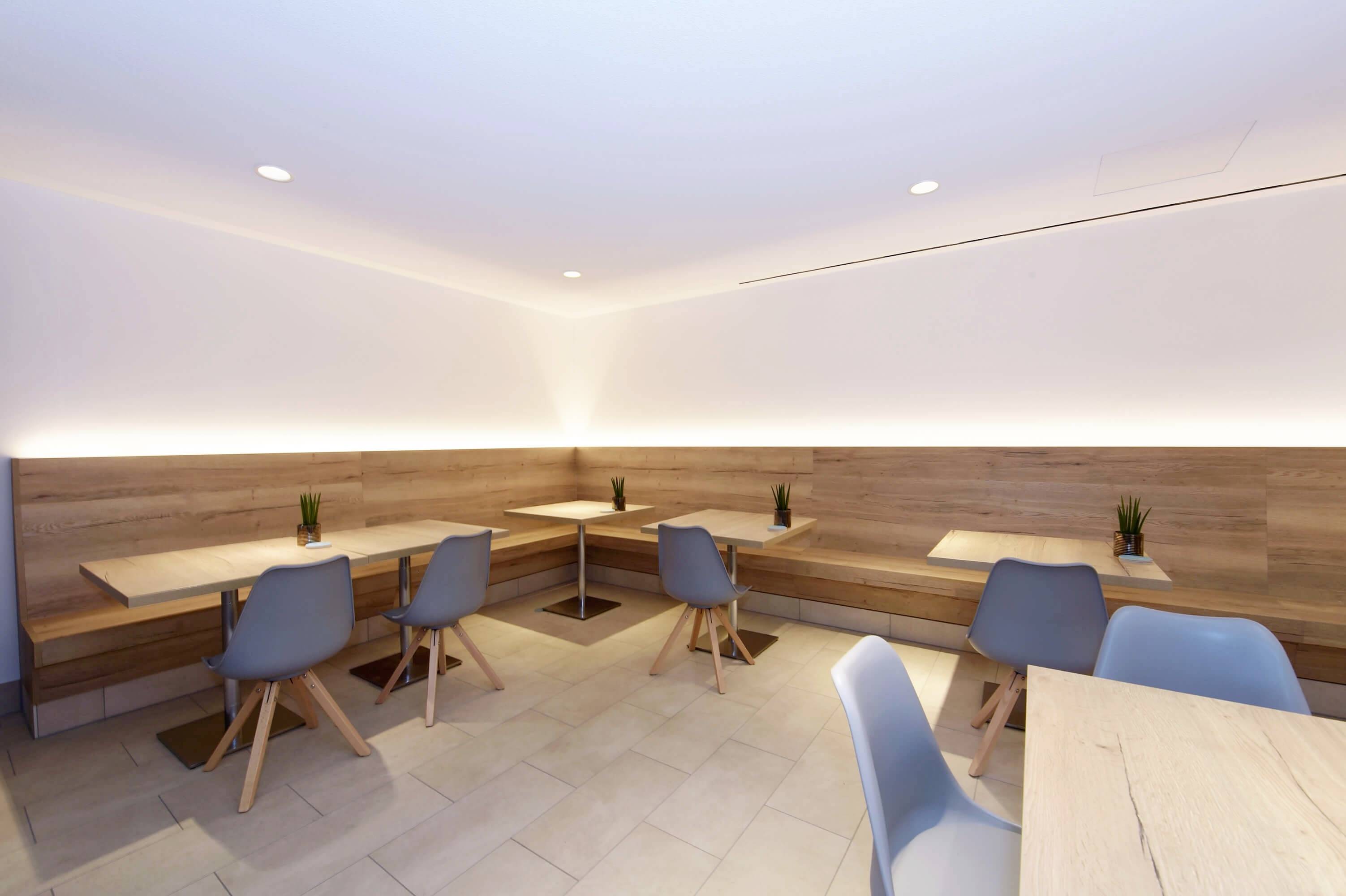 Moderná jedáleň s drevenými stolmi a sivými stoličkami. LED osvetlenie vytvára príjemnú atmosféru. Malé rastliny zdobia stoly.