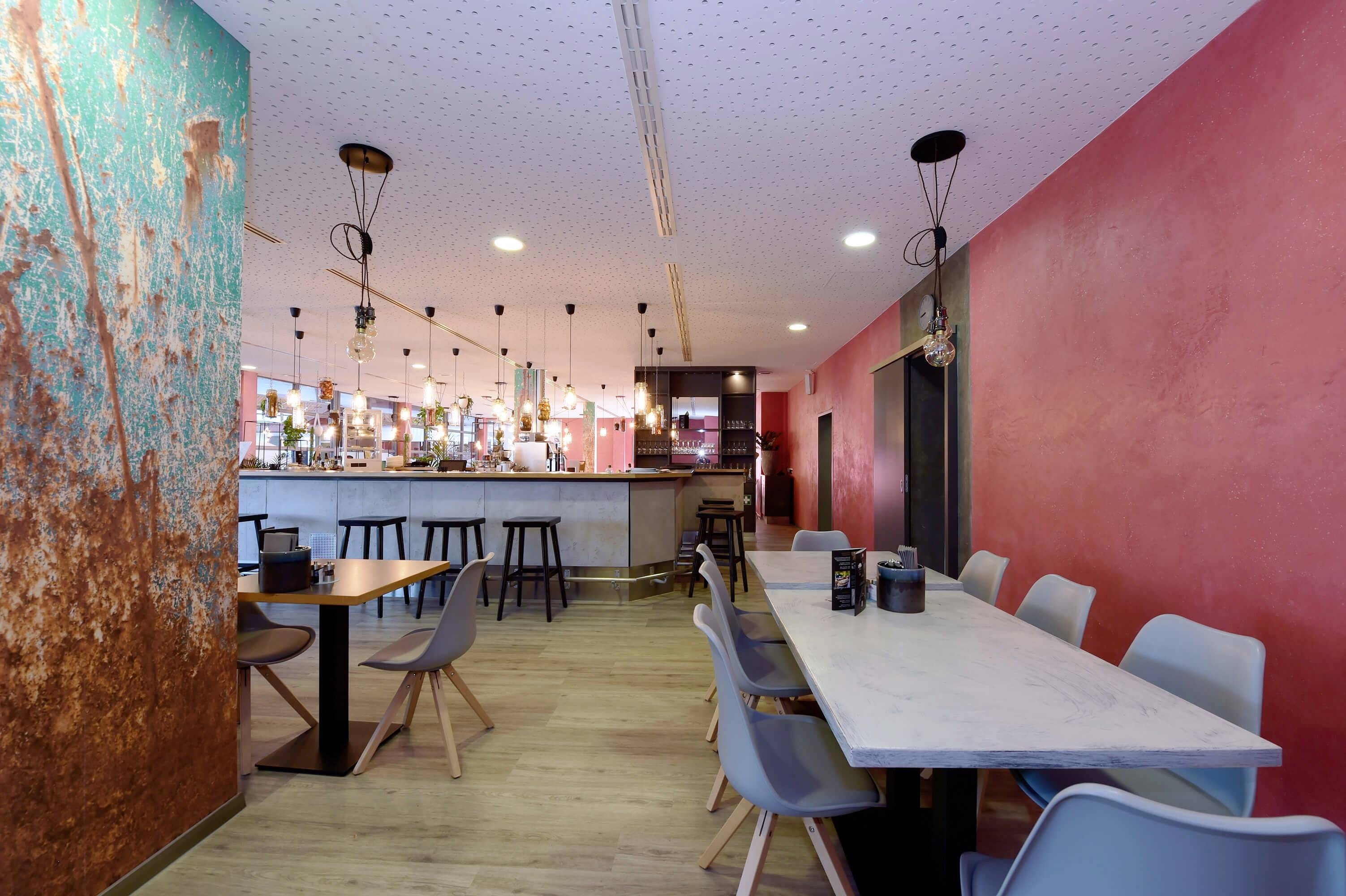 Moderná kaviareň s farebnými stenami, dlhým barovým pultom, sedacími priestormi s sivými stoličkami, štýlovým osvetlením a drevenou podlahou.