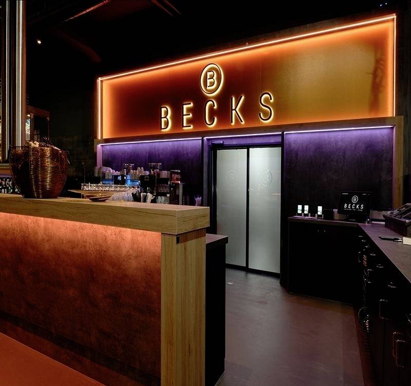 Bar s jasným, oranžovým neónovým nápisom 'BECKS' nad vstupom. Tmavý interiér, nápojový bar vľavo, moderná atmosféra.