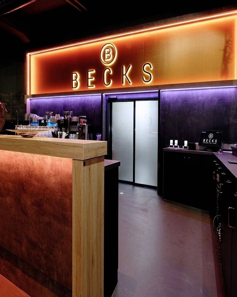 Štýlovo osvetlený vstupný priestor so svietiacim nápisom 'Becks'. Moderný, tmavý interiér s barovým pultom a dekoráciami.