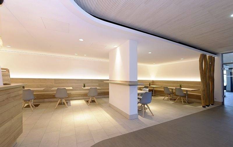 Moderný, minimalisticky navrhnutý kavirenský priestor so drevenými stolmi a sivými stoličkami, obklopený teplým nepriamym osvetlením.