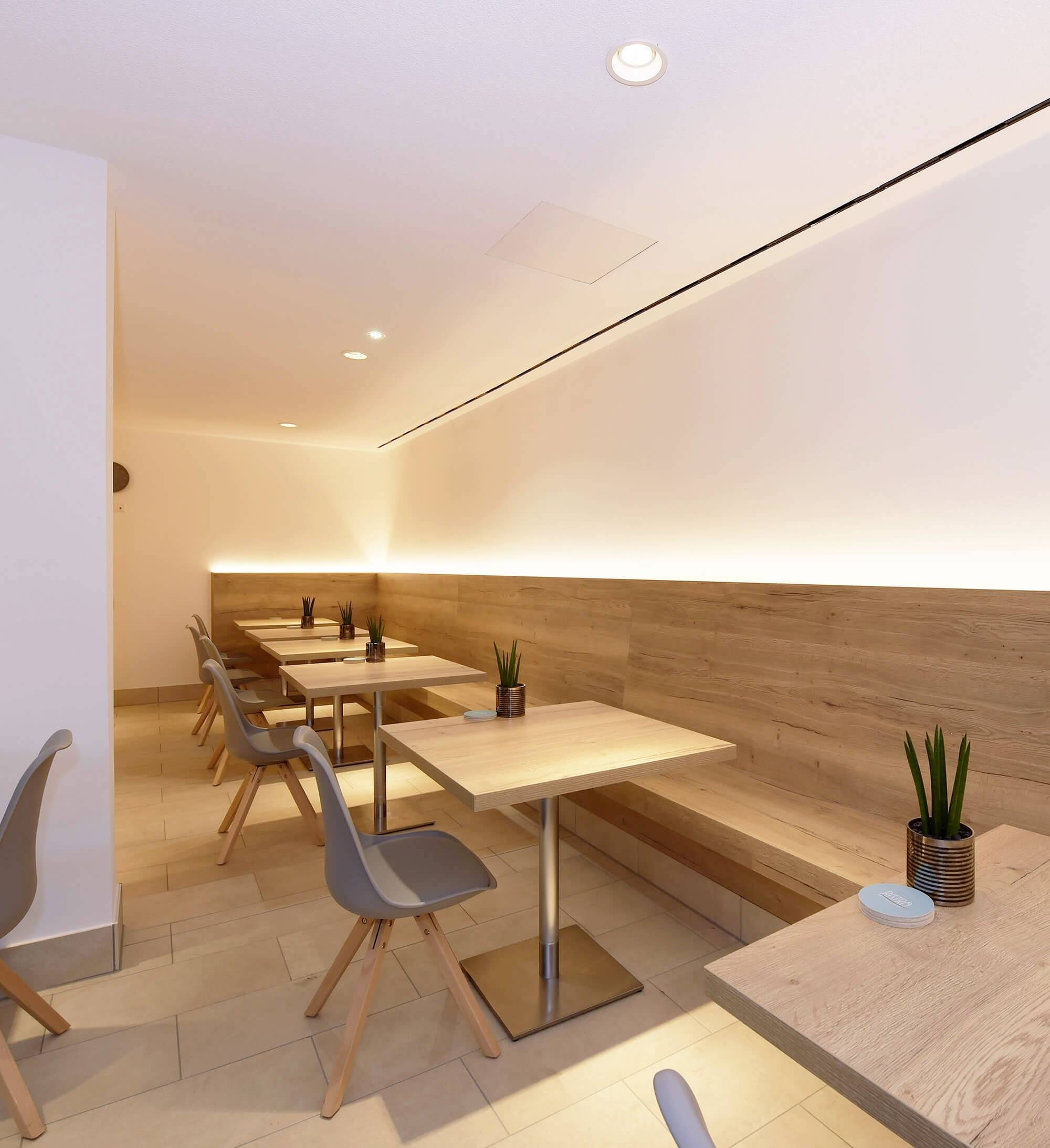 Moderná kaviareň s drevenými stolmi a sivými stoličkami, jemné svetlo na stene, príjemná a minimalistická atmosféra.