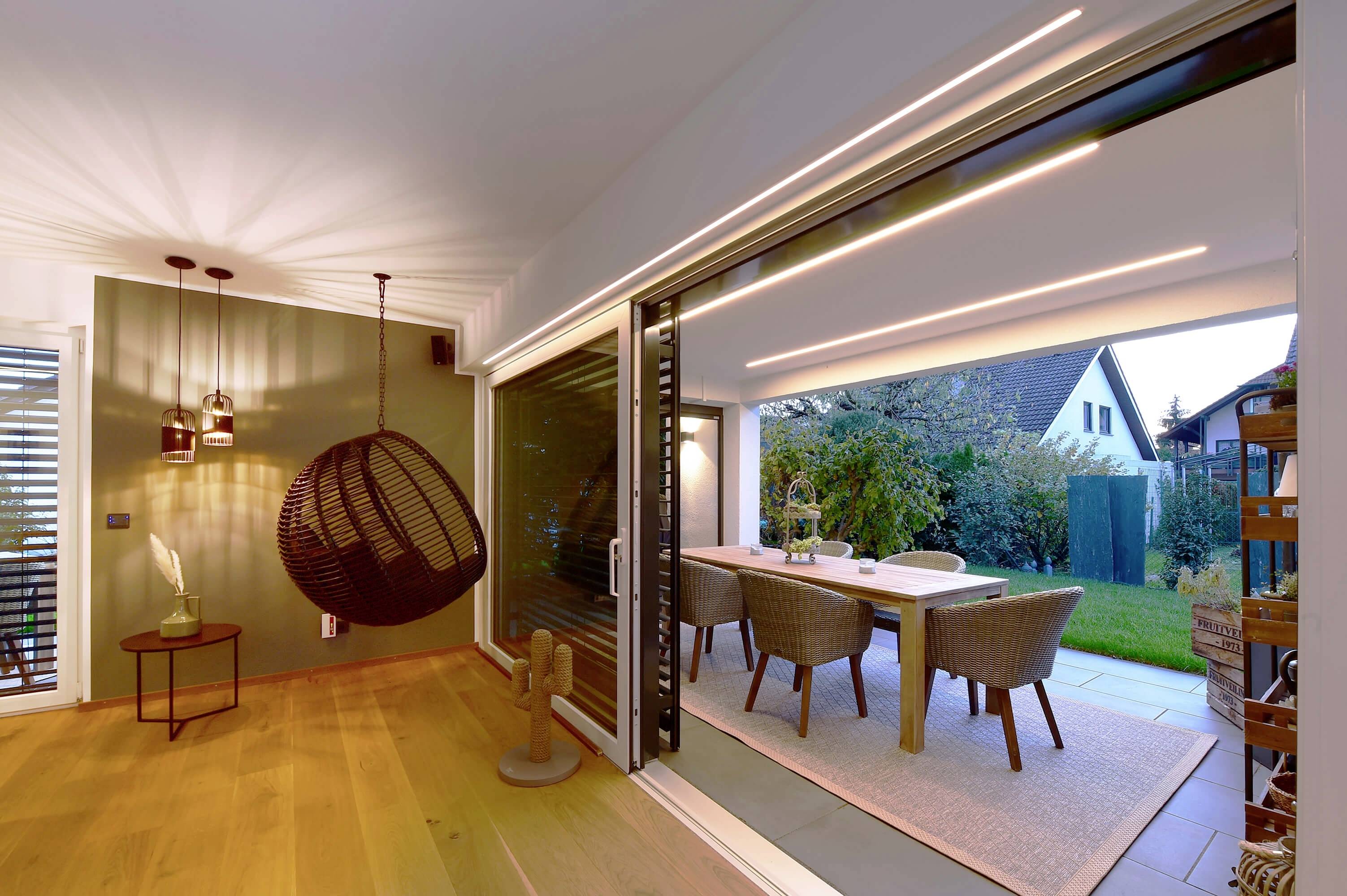 Moderná obývačka s závesným kreslom sa otvára na terasu s jedálenským stolom, obklopená záhradou a osvetlenou oblohou.