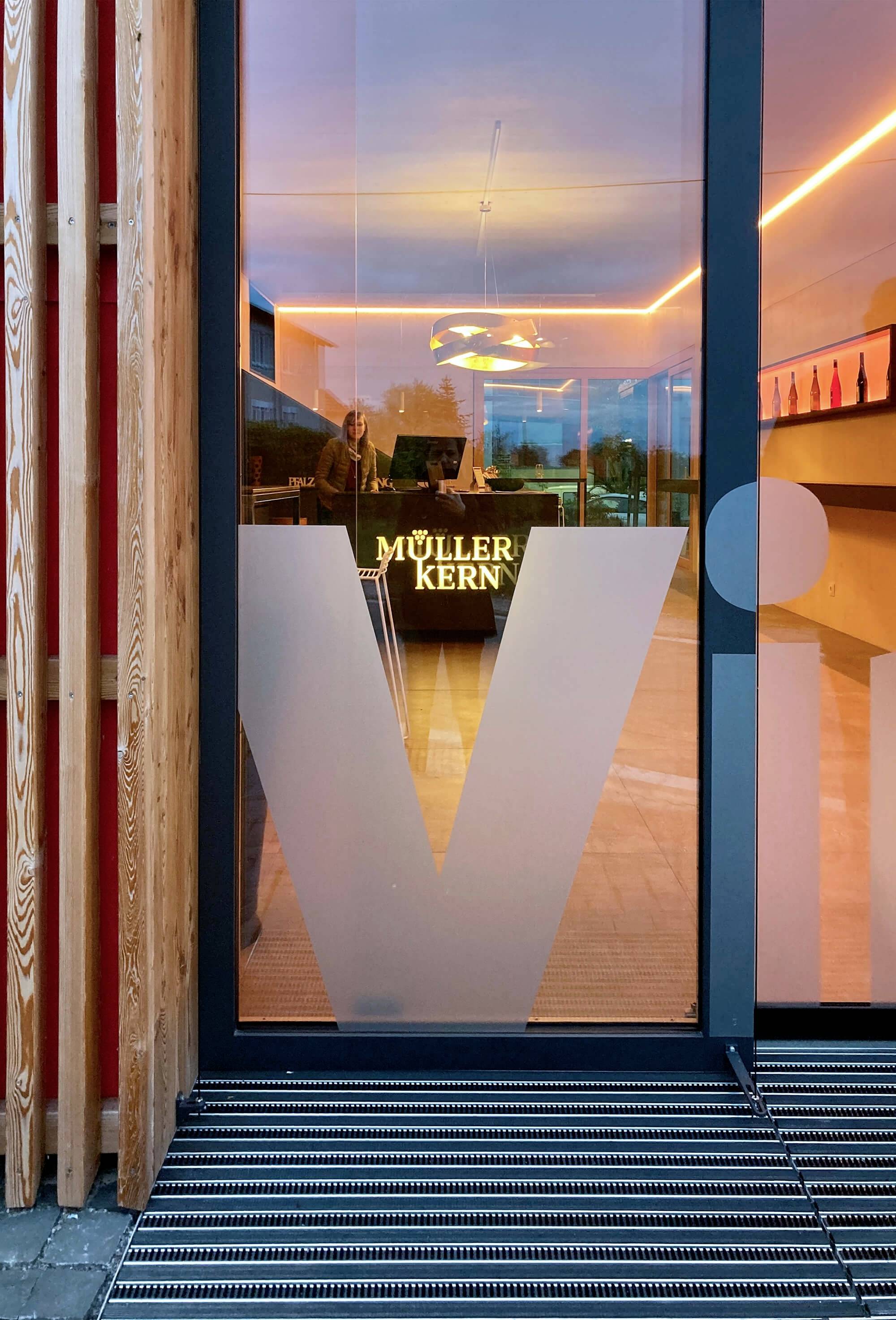 Sklenené okno s nápisom 'Müller Kern', za ním osvetlený interiér s drevenými prvkami a viditeľnými dvoma osobami.