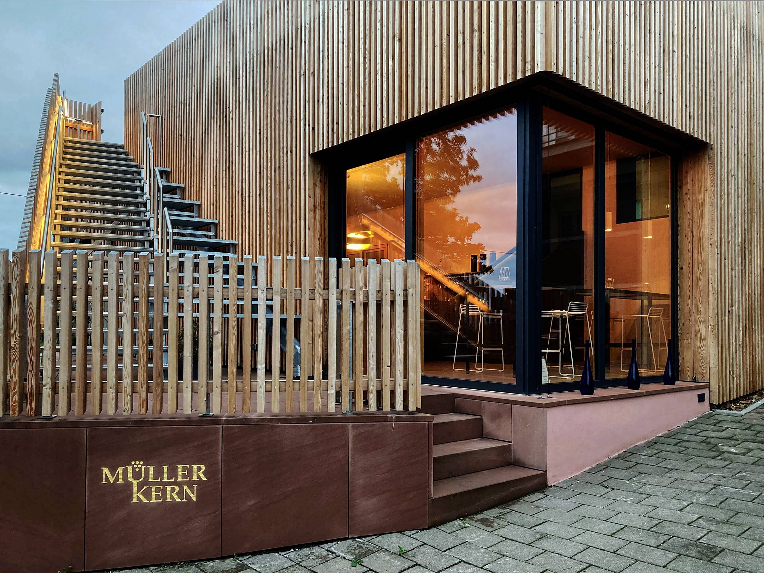 Moderná budova s drevenou fasádou a veľkými presklenými oknami, schodisko na ľavej strane, v popredí logo 'Müller Kern'.