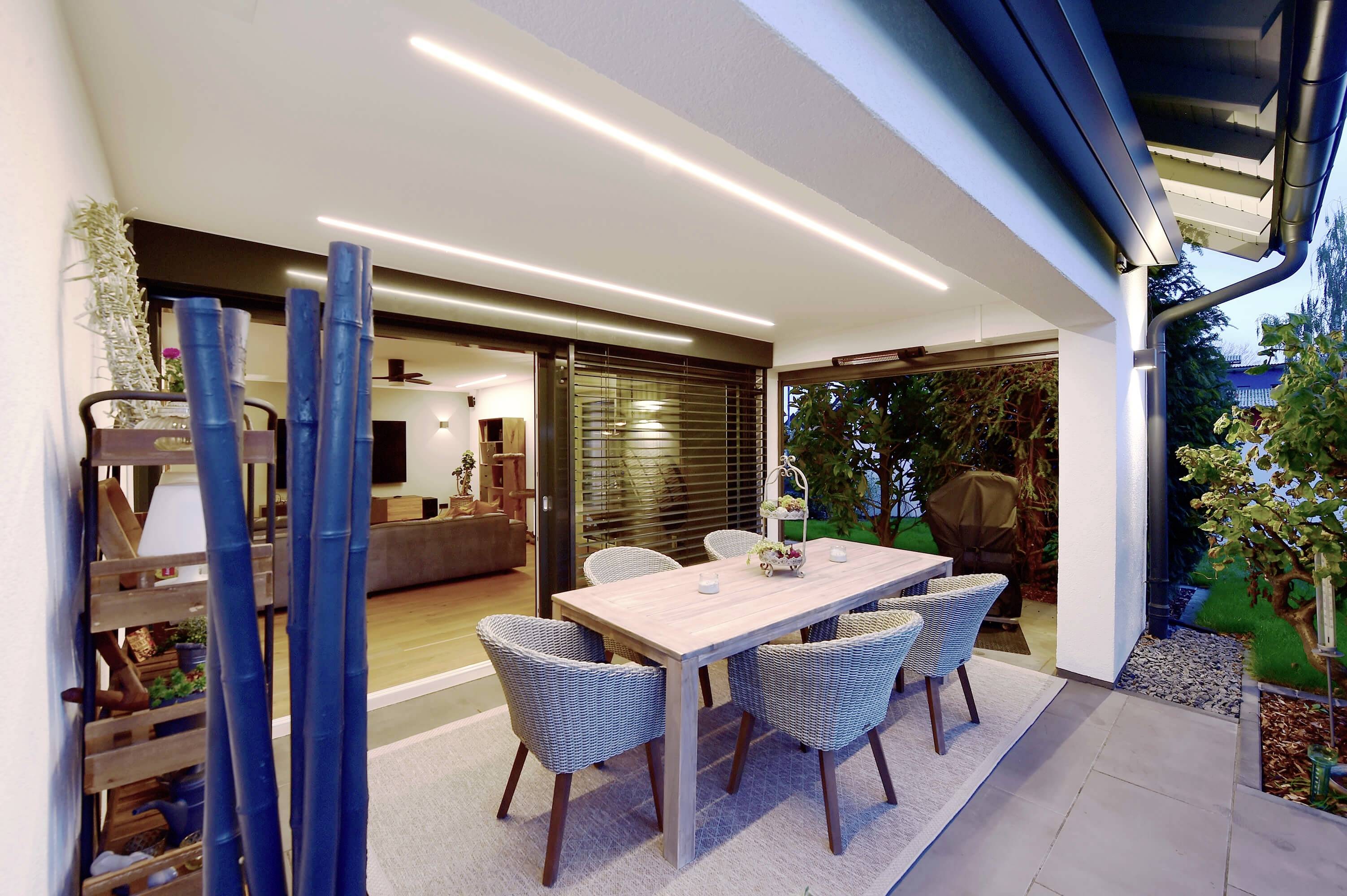 Zakrytá terasa s dreveným stolom a šiestimi stoličkami, otvorená do moderného obývacie priestoru. Záhrada v pozadí.