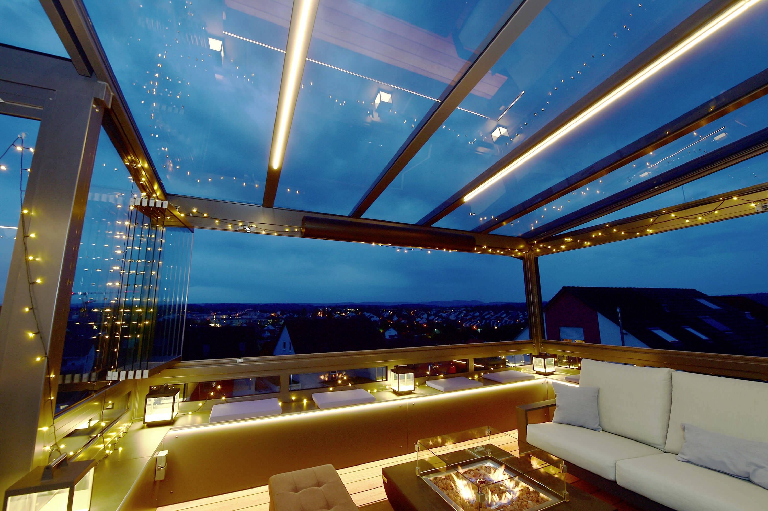 Zakrytá terasa v noci s LED svetelnými reťazami, sedačkou a ohňovou misou. Výhľad na mesto v pozadí pod tmavou oblohou.