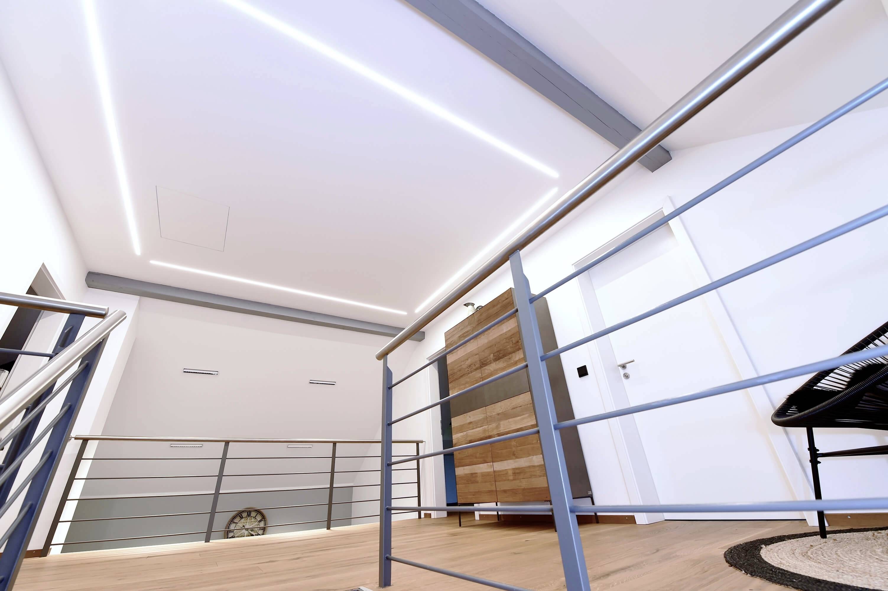 Moderná chodba s drevenou podlahou, kovovým zábradlím a akcentným osvetlením, vedúca k bielym dverám. Atmosféra je minimalistická a čistá.