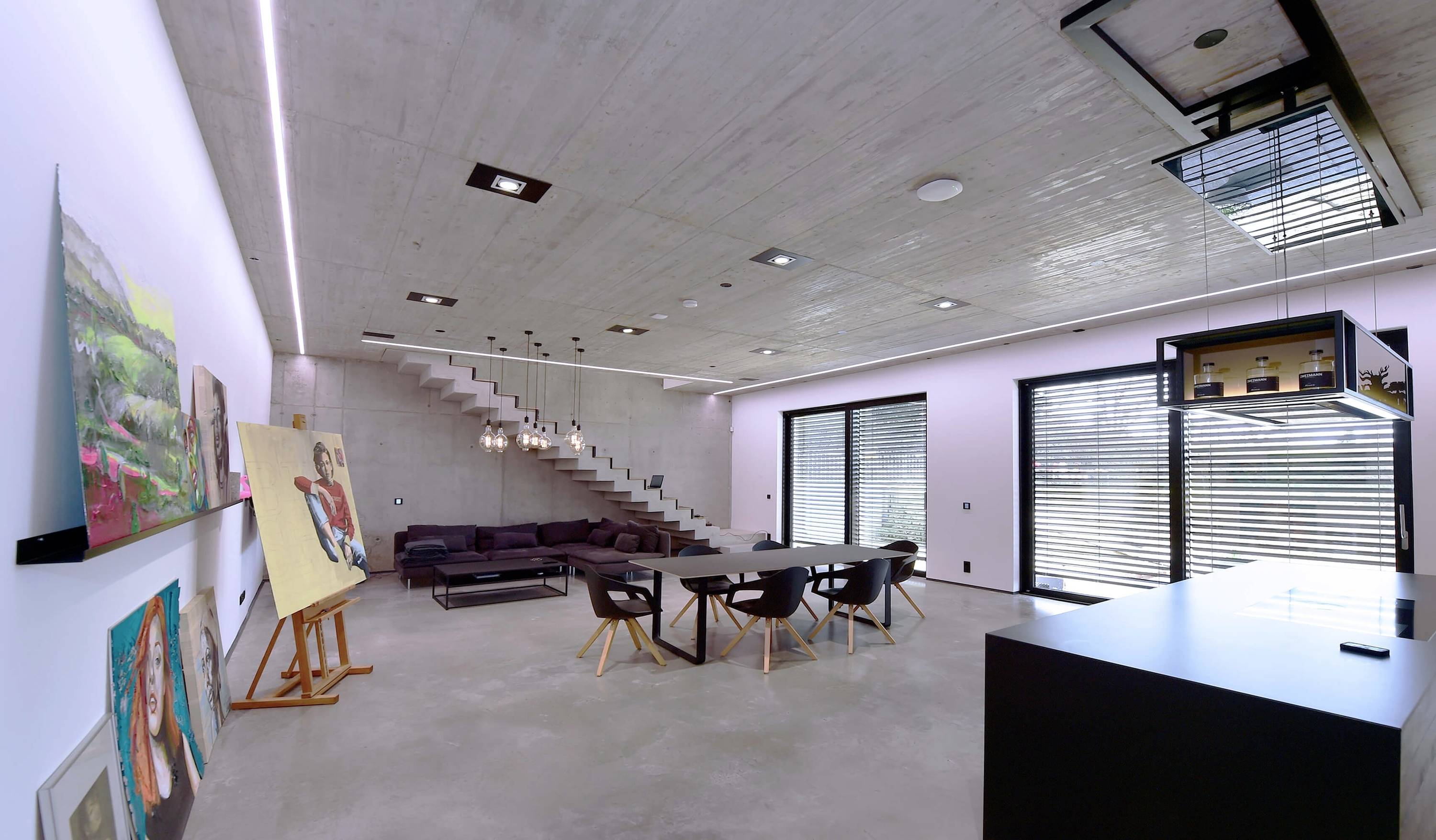Moderný obytný priestor s betónovými stenami a stropom. Otvorená dispozícia s veľkým stolom, stoličkami, sedačkou a schodiskom. Umelecké diela na stenách.