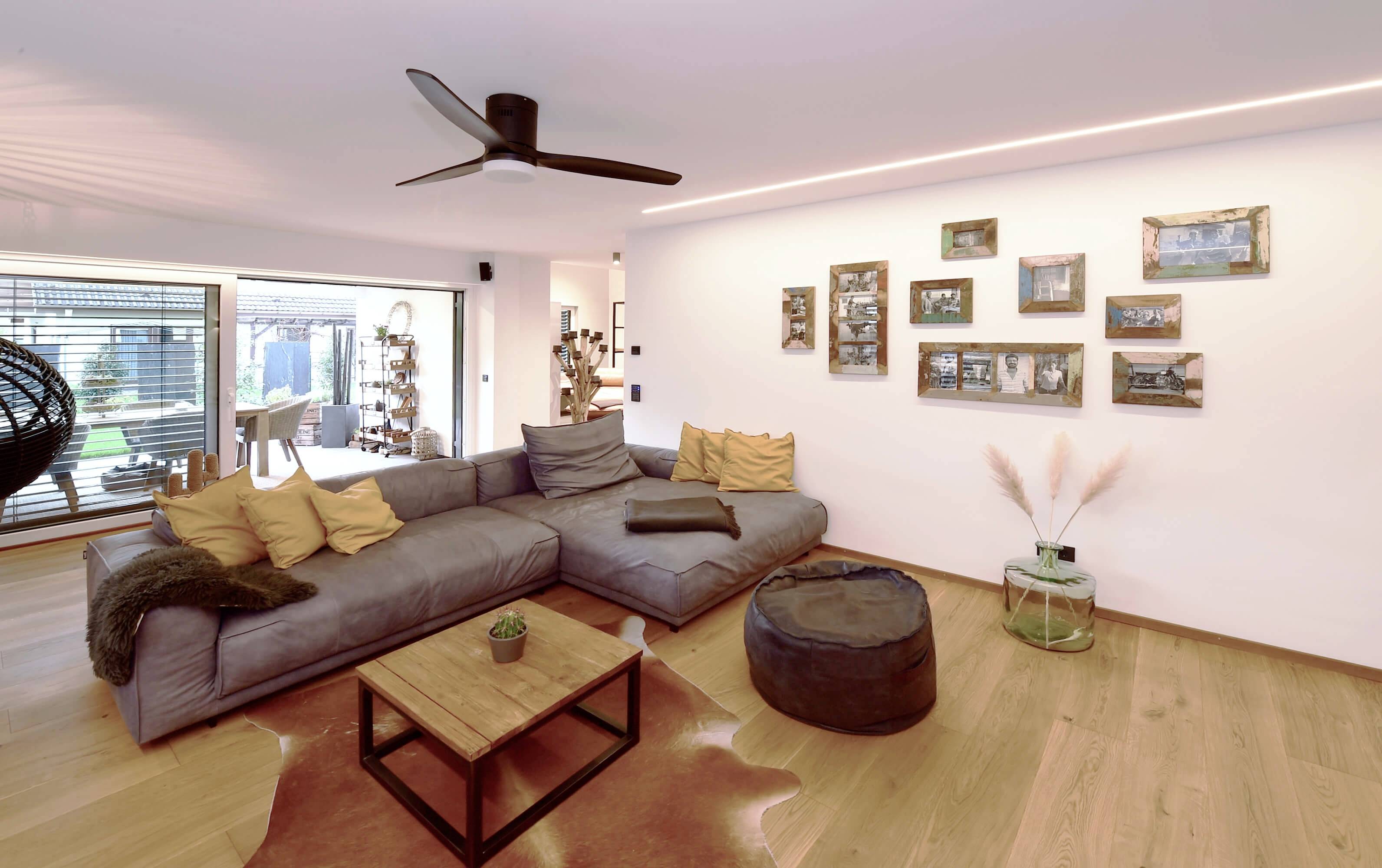 Veľká obývačka so sivou sedačkou, čiernym stropným ventilátorom, rámikmi na stenách a vstupom do slnečnej záhrady.