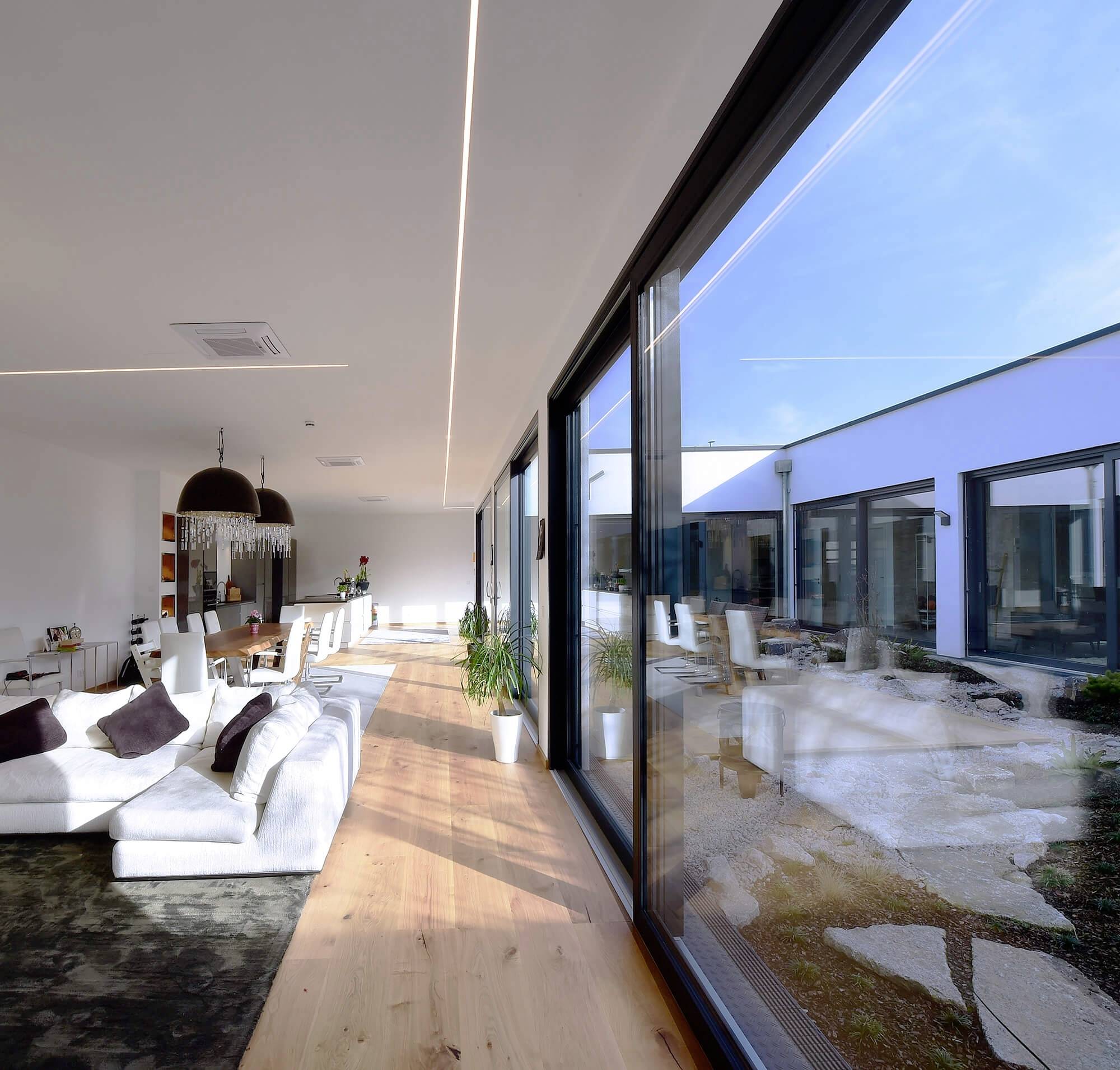 Moderný obytný priestor s veľkými presklenými oknami, ktoré ponúkajú výhľad na slnečný vnútorný dvor. Svetlý nábytok, minimalistický dizajn.