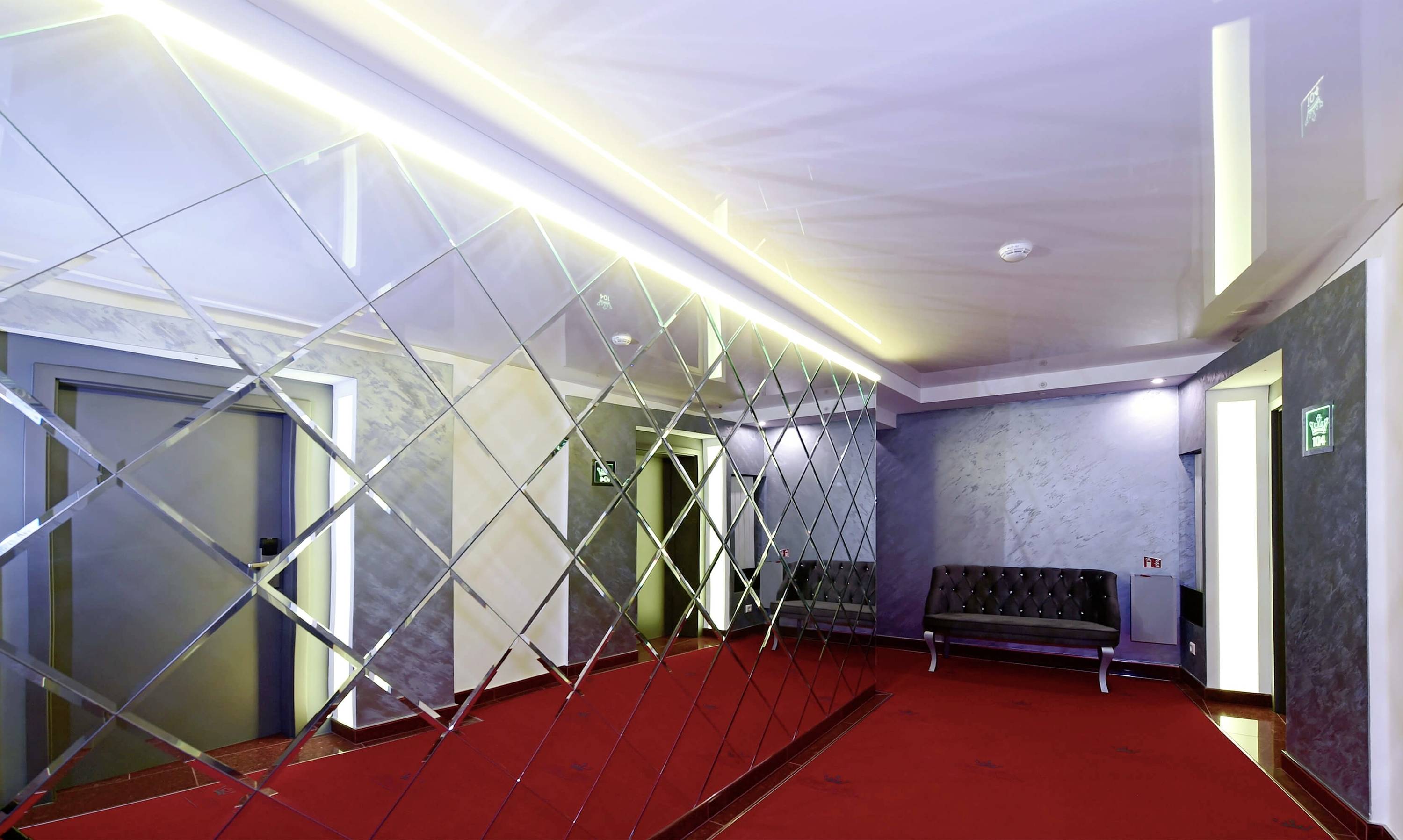 Chodba s červeným kobercom, moderným osvetlením, striebornými stenami a veľkým zrkadlom. V pozadí sú viditeľné dvere a lavica.