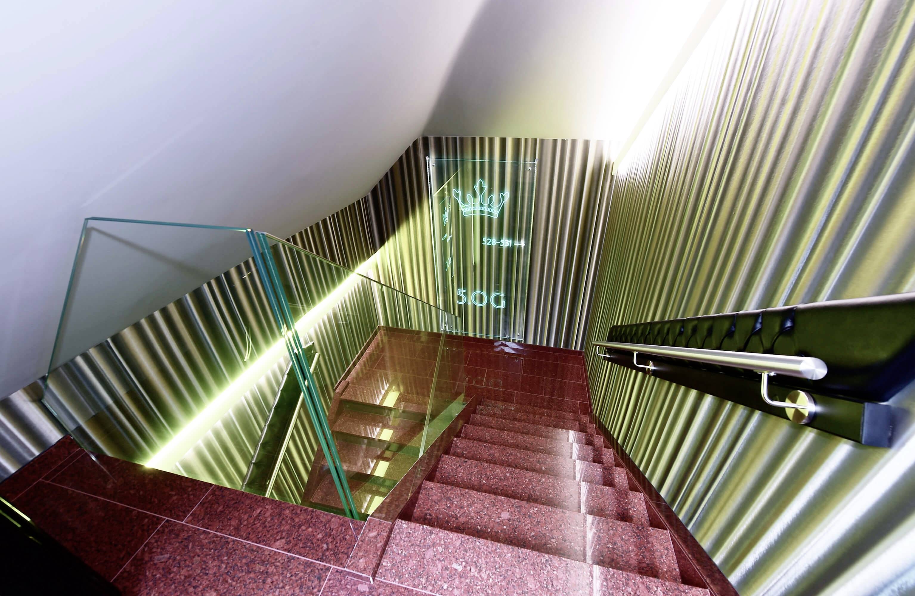 Červené schodisko v modernej budove so skleneným zábradlím a kovovými stenami. Na stene je premietaná koruna a číslo '500'.
