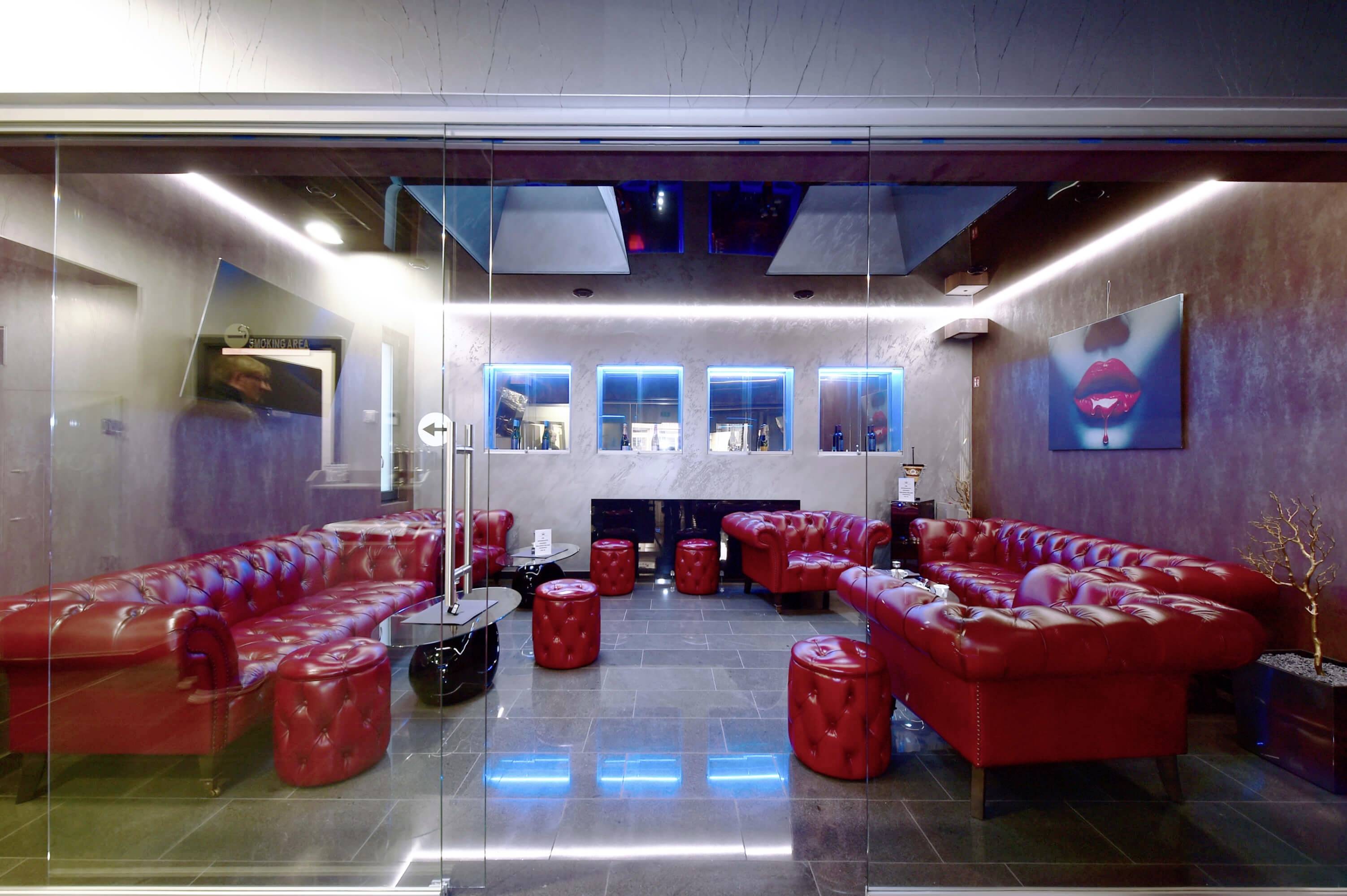 Červená kožená sedacia oblasť v modernej salónke s neónovo modrým osvetlením, abstraktnými nástennými maľbami a skleneným stolom.