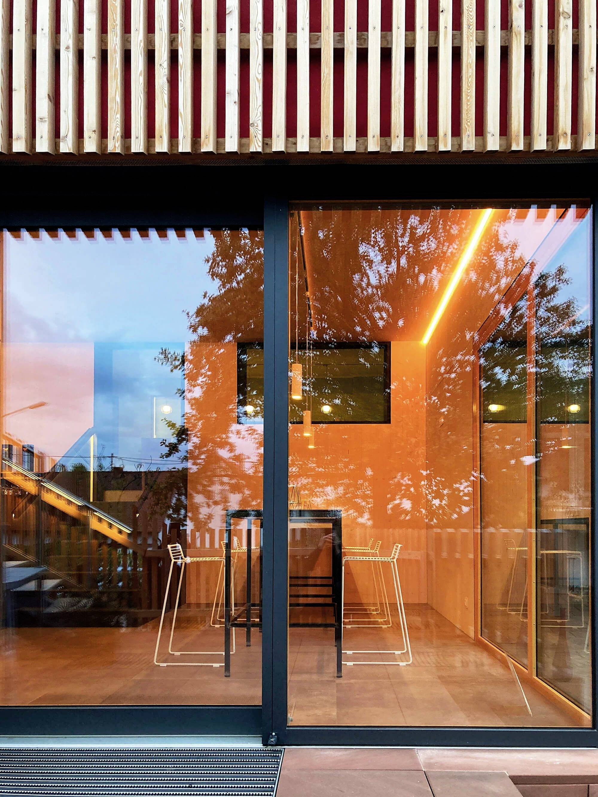 Moderná sklená fasáda budovy s oranžovým osvetlením. Vnútri sú viditeľné stoly a stoličky, stromy sa odrážajú v skle.