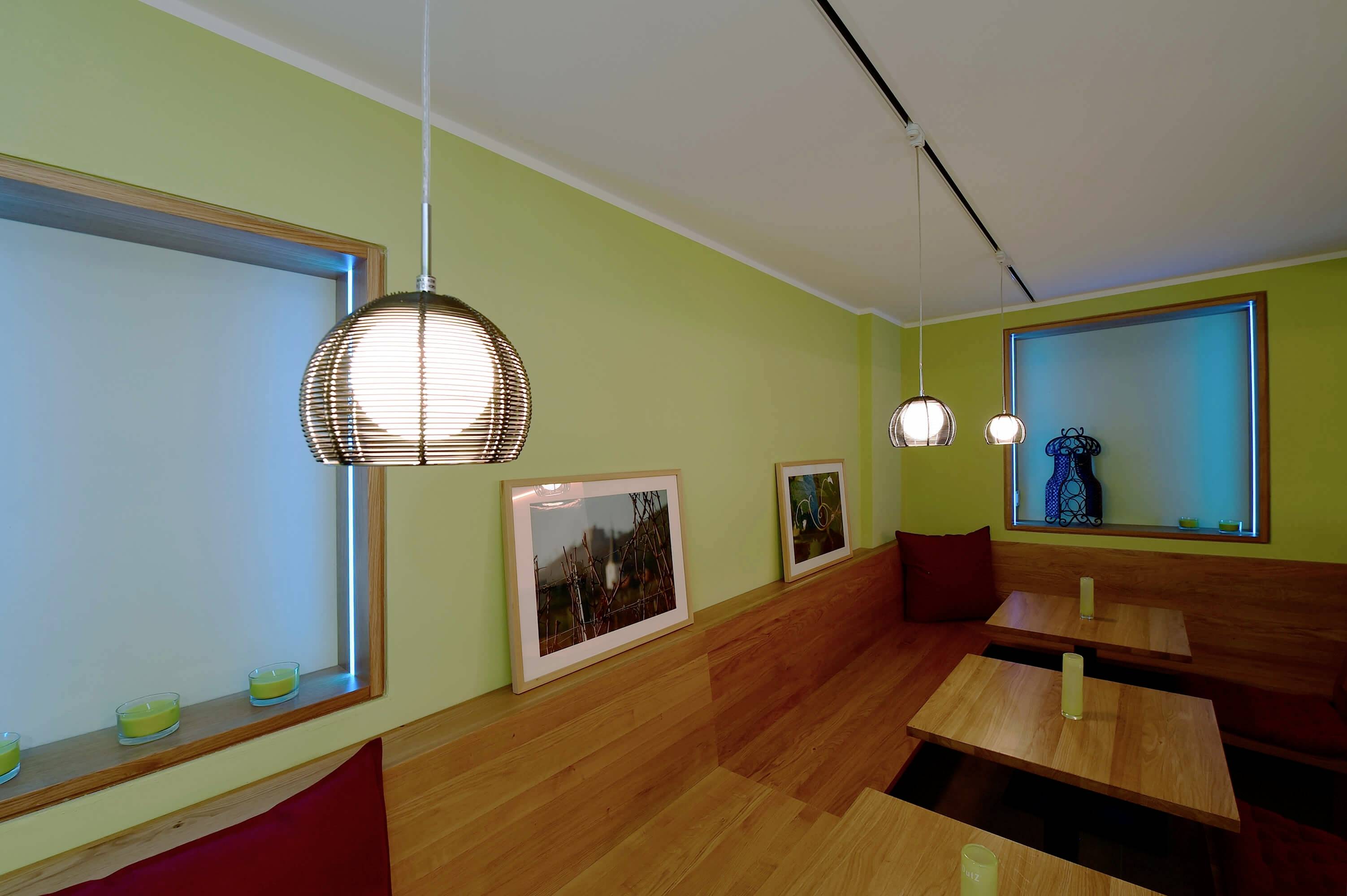 Malá kaviareň s drevenými sedadlami, zelenými stenami a visiacimi lampami. Veľké okná prepúšťajú veľa svetla dovnútra.