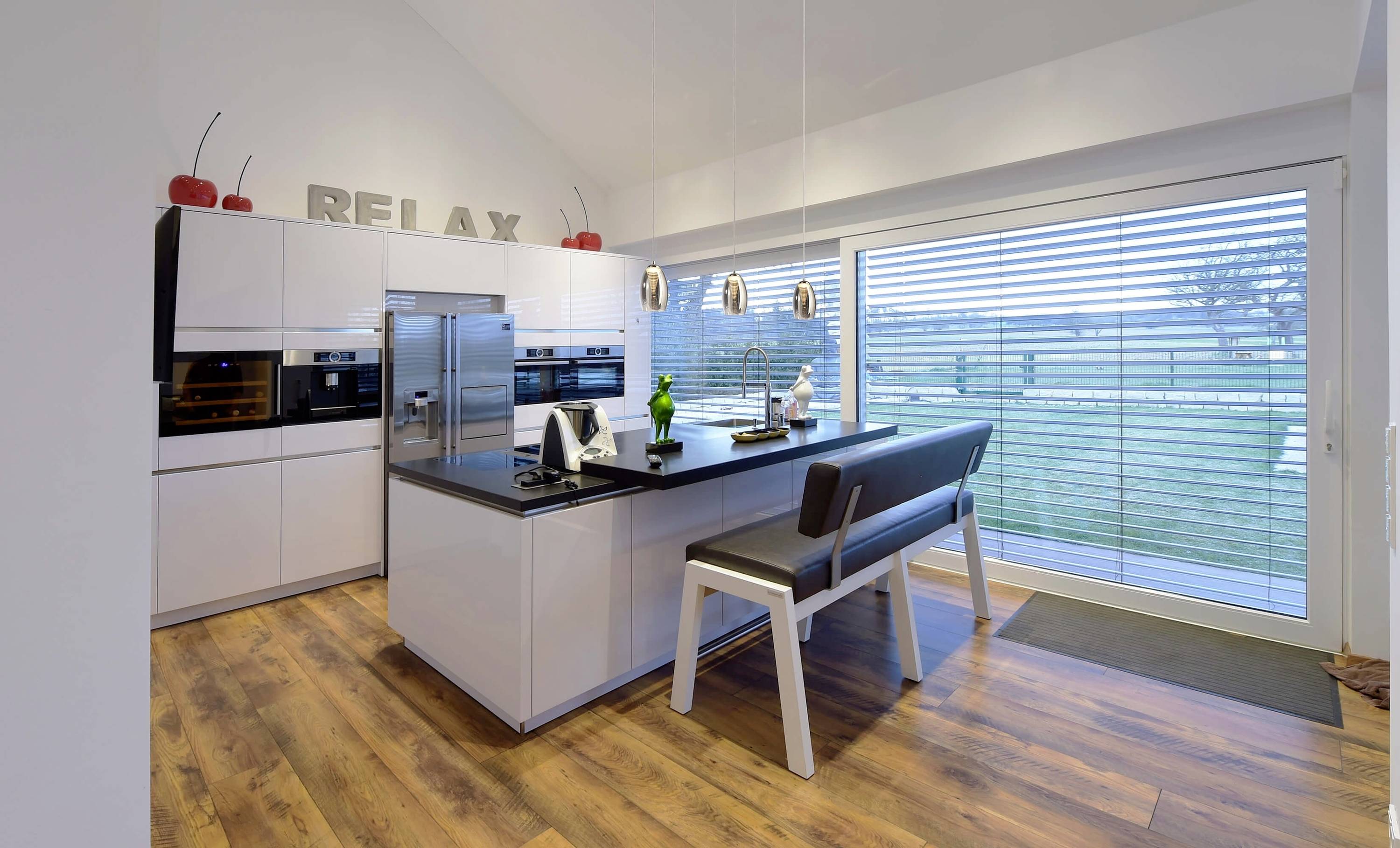 Moderná kuchyňa s kuchynským ostrovčekom a integrovanými spotrebičmi. Veľká presklená stena ponúka výhľad do zelene. Nápis 'RELAX' nad skrinkami.