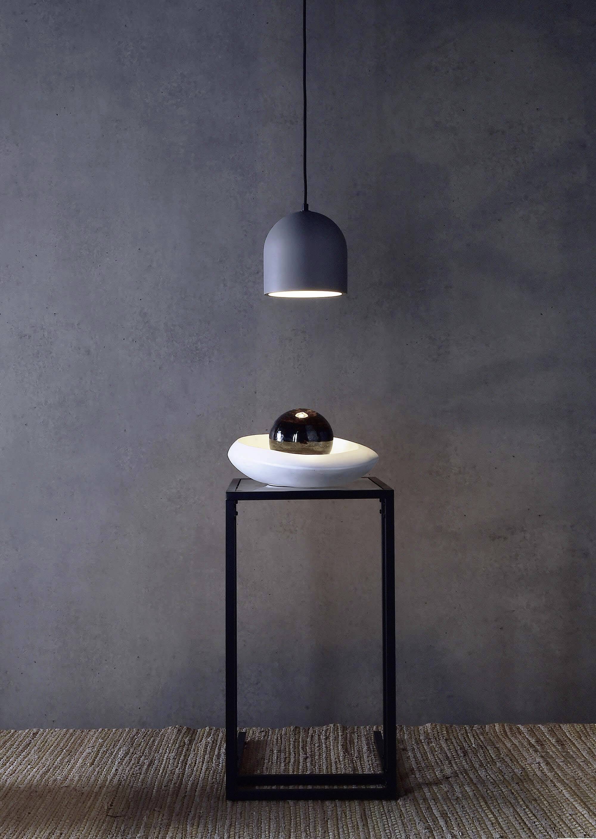 Minimalistická miestnosť so zavesením lampou nad modernou sochou umiestnenou na úzkom stolíku pred sivou stenou.