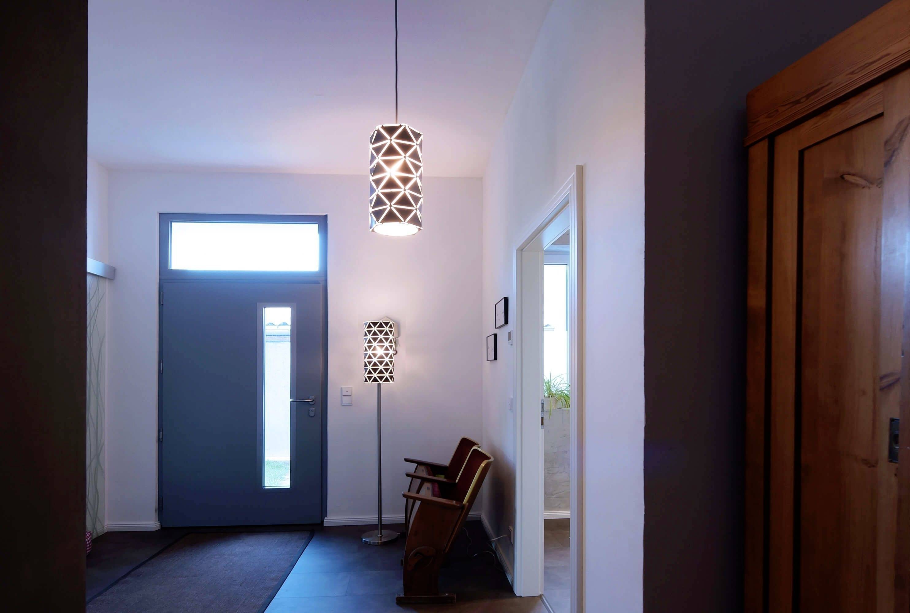 Vstupný priestor domu s modrou bránou, dvoma lampami a dvomi stoličkami pred bielou stenou, pôsobí minimalisticky a moderne.