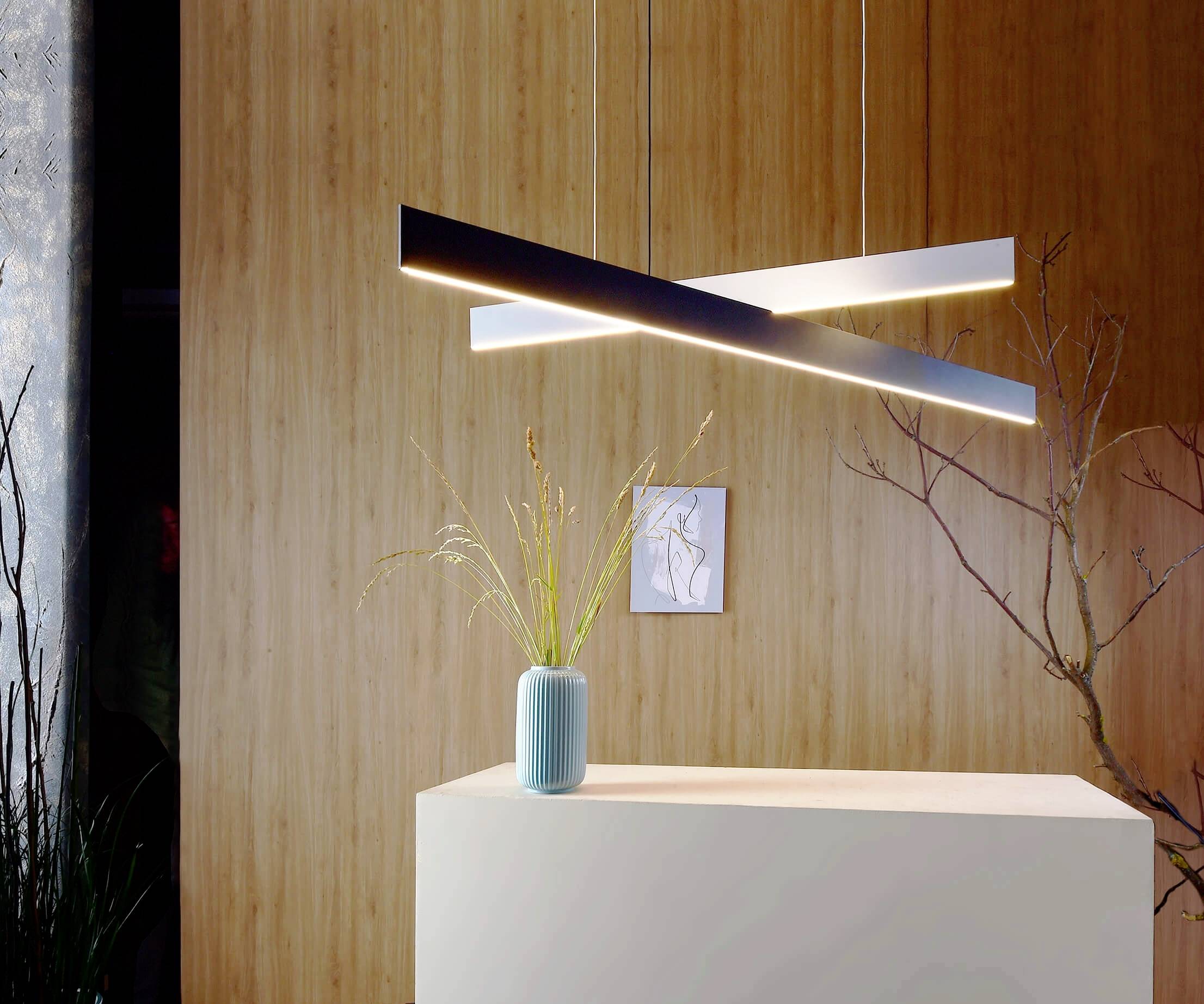 Moderná, minimalistická závesná lampa s dvoma križujúcimi sa svetelnými tyčami; pod ňou stojí zelená váza s trávami.