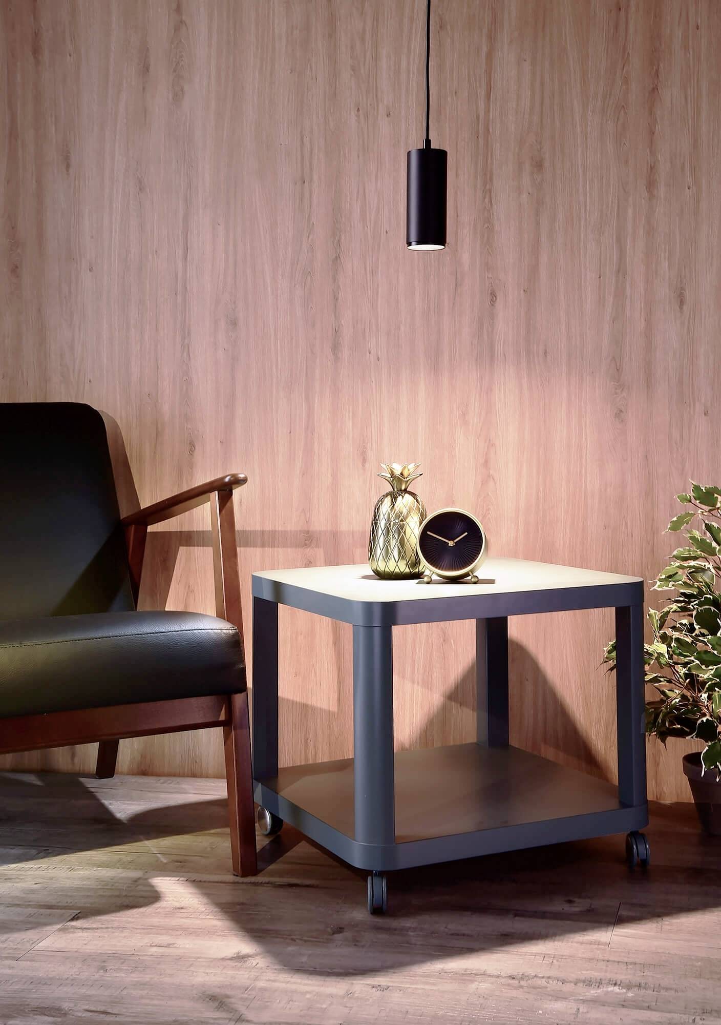 Malý drevený stolík s čiernym hodinami a zlatou vázou, vedľa hnedé kožené kreslo a závesná lampa, vpravo rastlina.