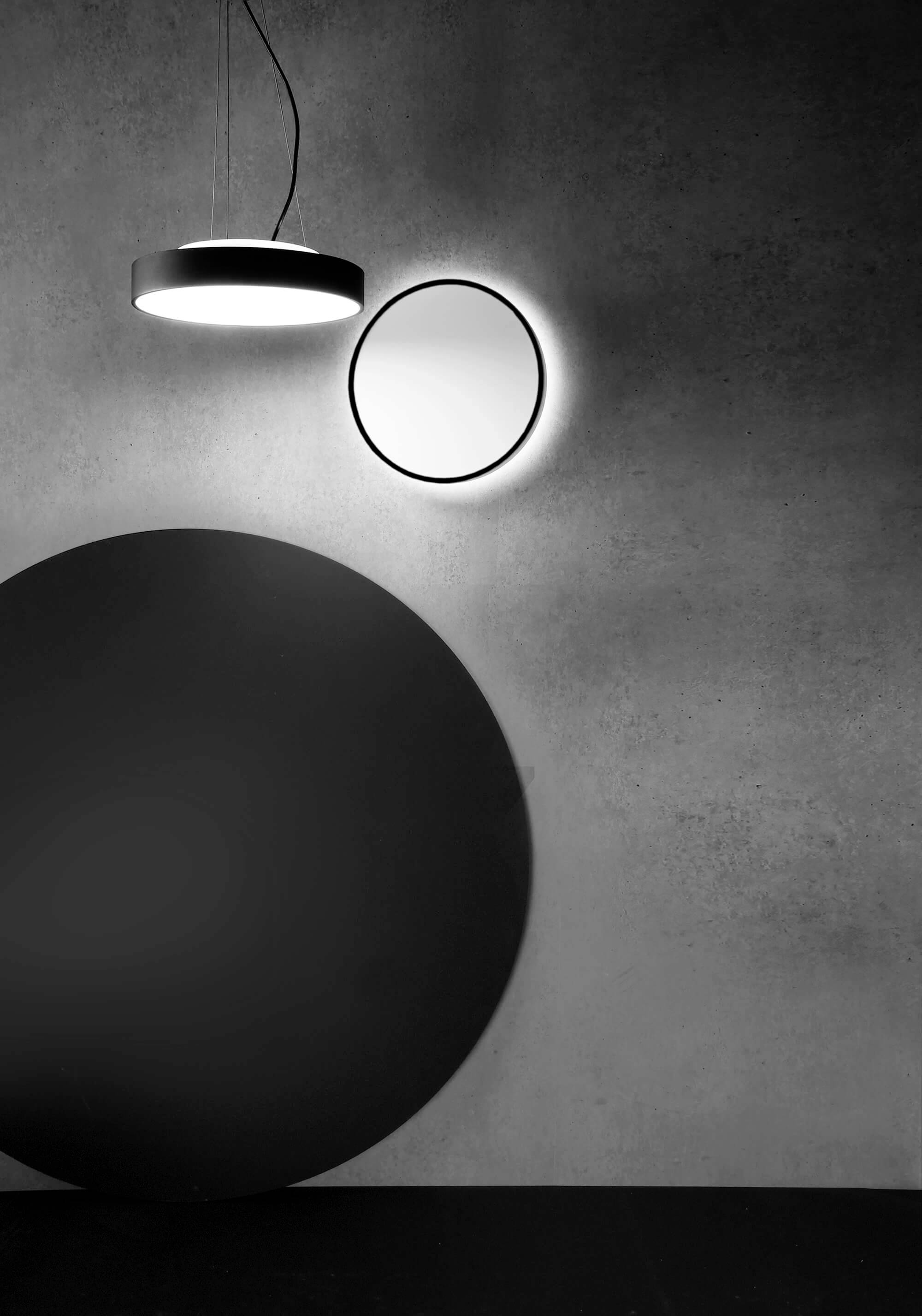 Čiernobiela fotografia s moderným stropným svietidlom, ktoré vytvára guľatý tieň na béžovej stene vedľa osvetleného zrkadla.