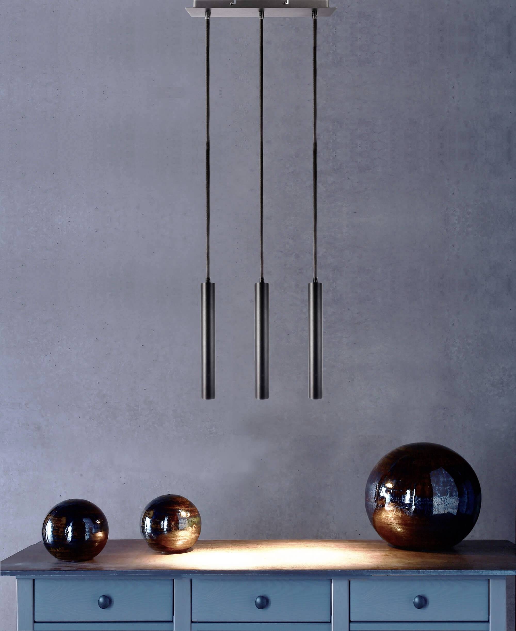 Závesná lampa s tromi valcovitými svetlami nad modrým stolom s tromi zrkadlovými guľami, ktoré zobrazujú svetelné odrazy.