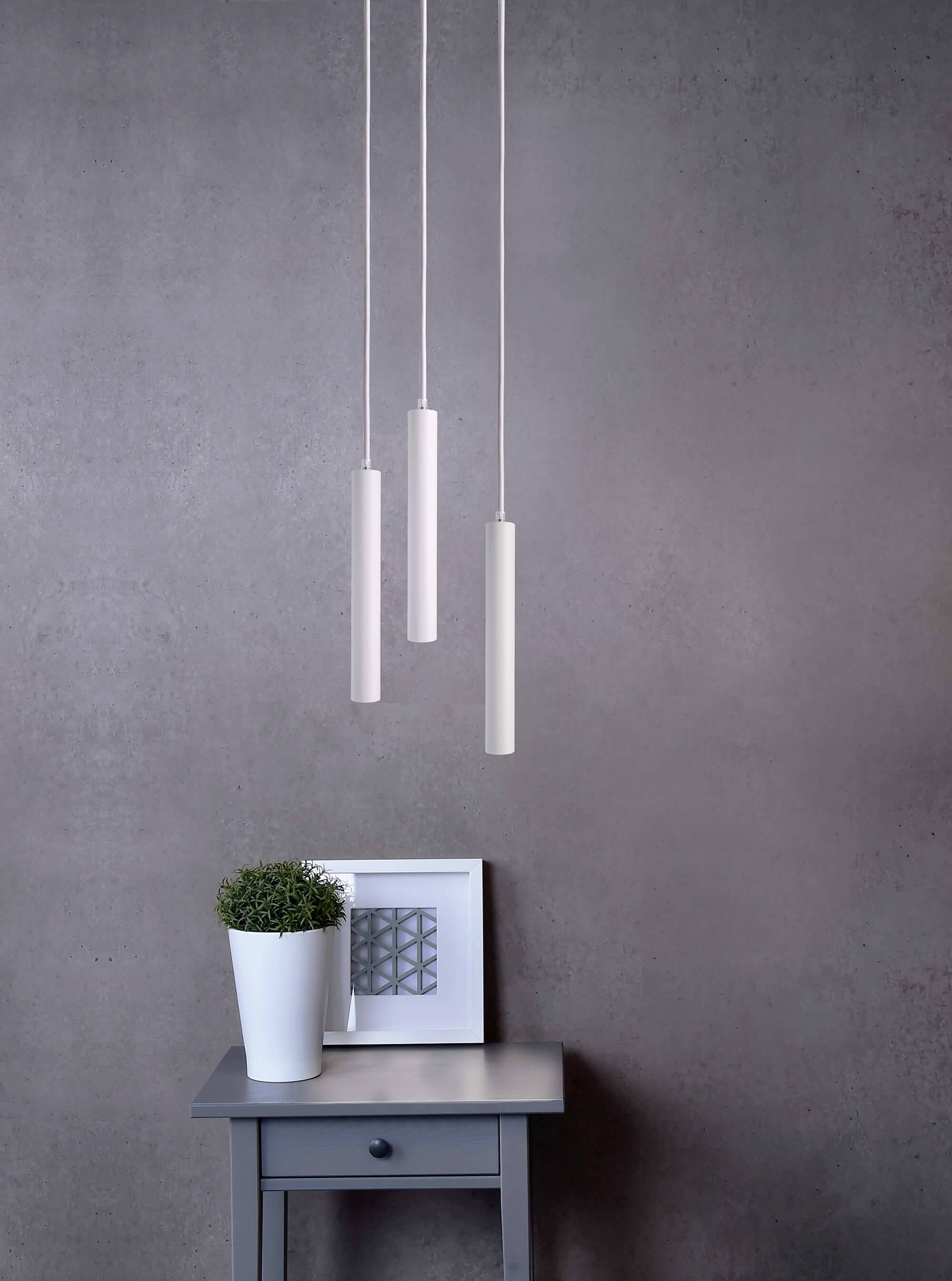 Minimalistické zariadenie izby s troma zavesenými valcovými lampami nad malým stolíkom, ktorý nesie rastlinu a zarámovaný obraz.