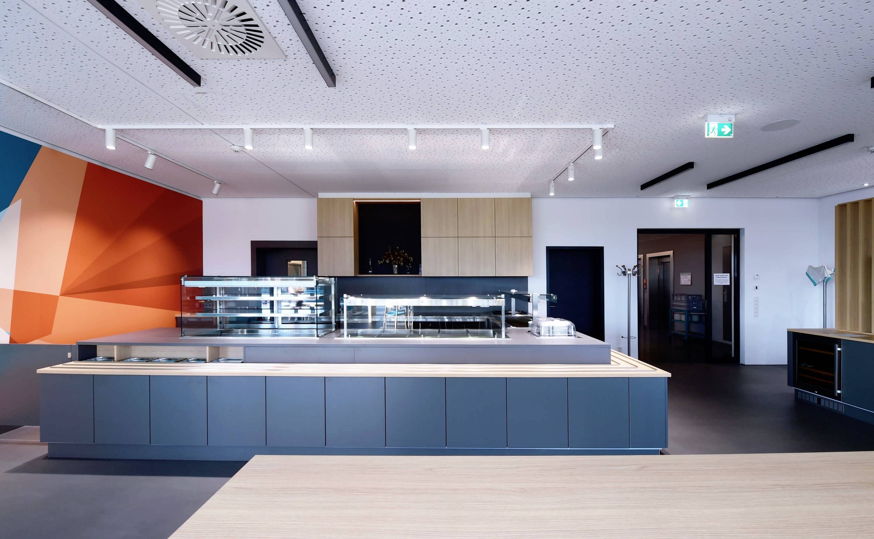 Moderná jedáleň s svetlomodrými pultmi, drevenými detailmi a pestrofarebnou stenou. Tlmené osvetlenie, žiadny personál ani hostia nie sú viditeľní.