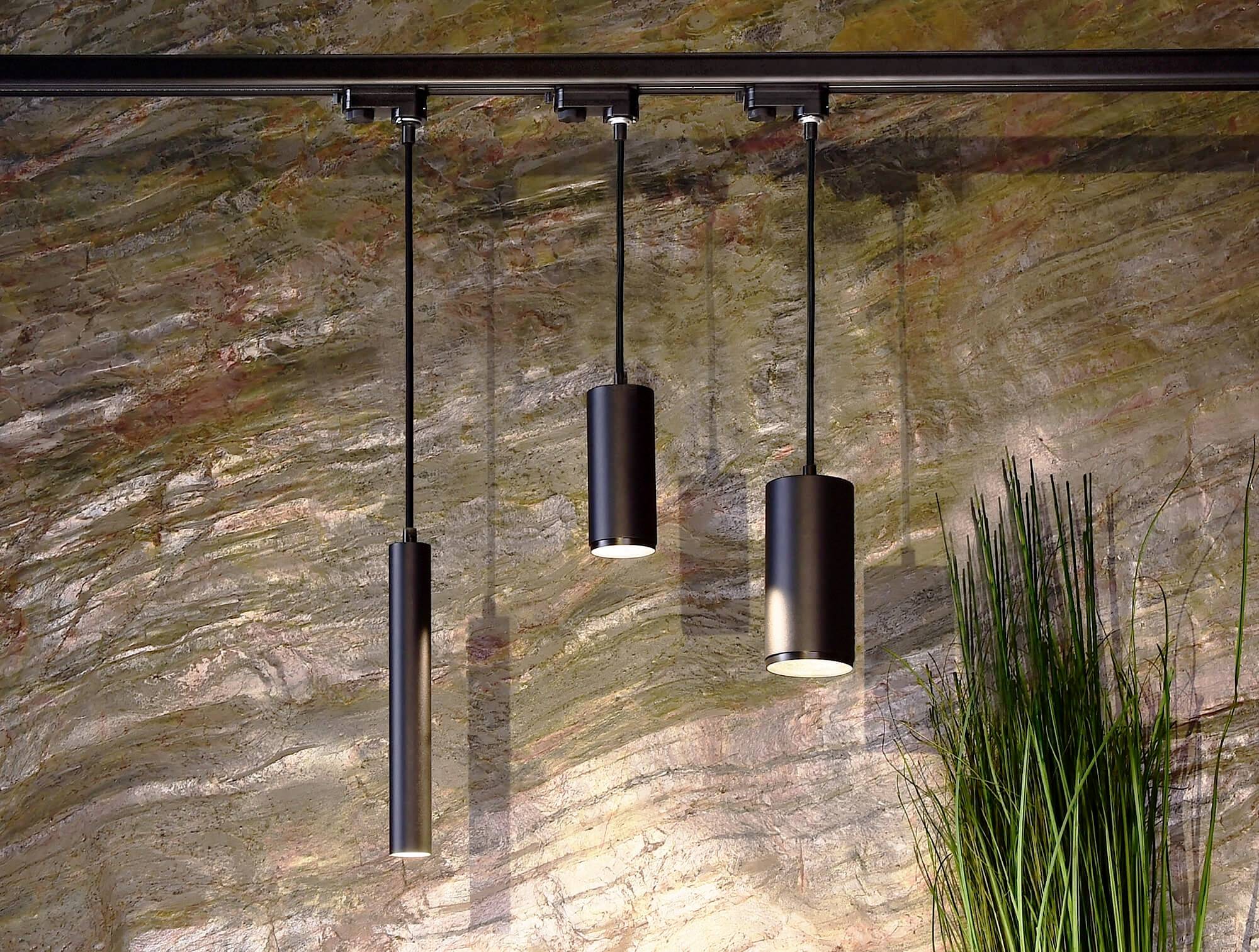 Tri čierne cylindrické závesné lampy pred štruktúrovanou kamennou stenou so <br> dekoratívnou trávou na pravej strane.