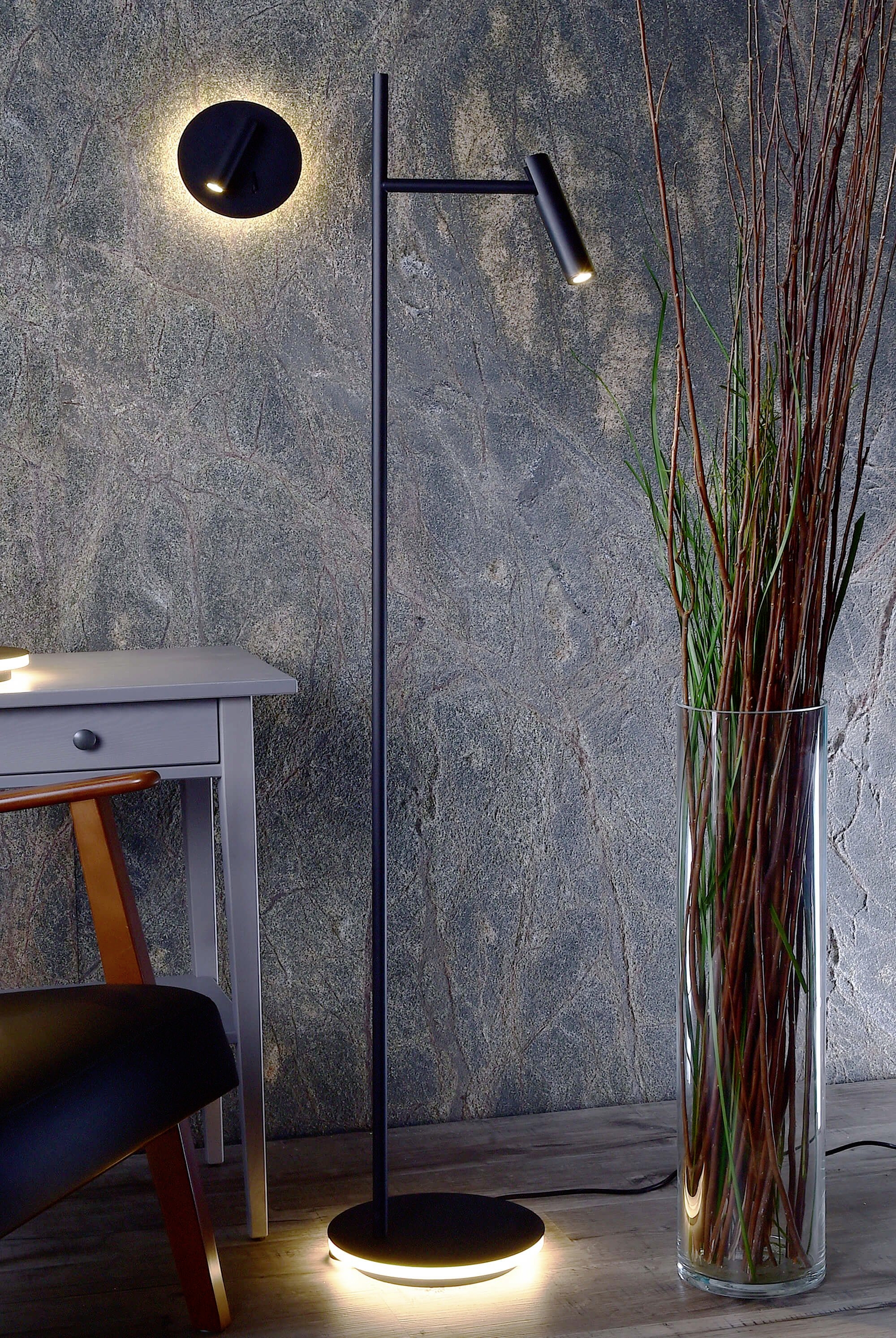 Stojacia lampa v modernom dizajne vedľa príručného stolíka, pred štruktúrovanou stenou. Vedľa nej vysoká váza s tenkými vetvami.