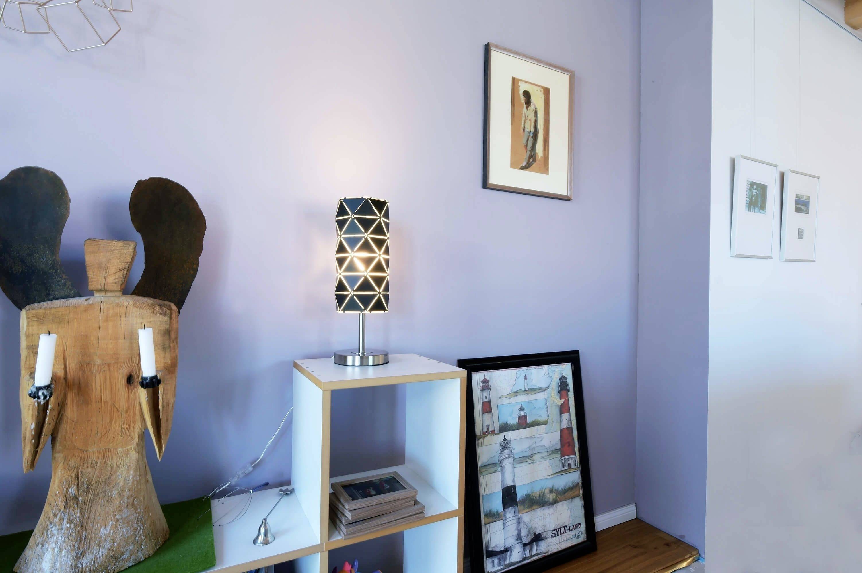 Miestnosť s fialovými stenami; drevená dekorácia na stene so sviečkovými stojanmi, police s dekoráciami, obraz na stene. Atmosféra je moderná a umelecká.