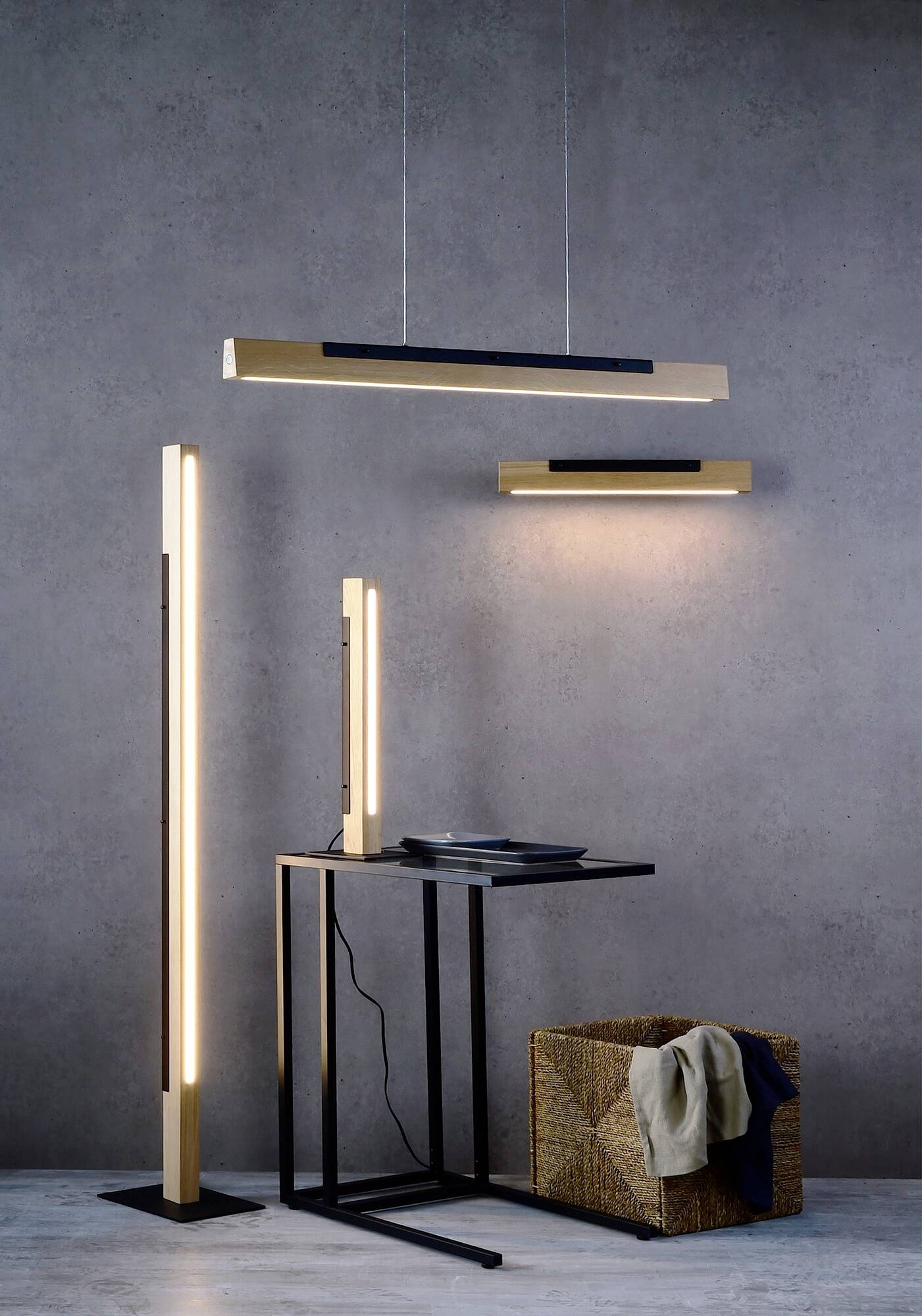 Drevená lampa v modernom dizajne vedľa malého stolíka s predmetmi. Svietidlá vyžarujú teplé svetlo na sivej stene.