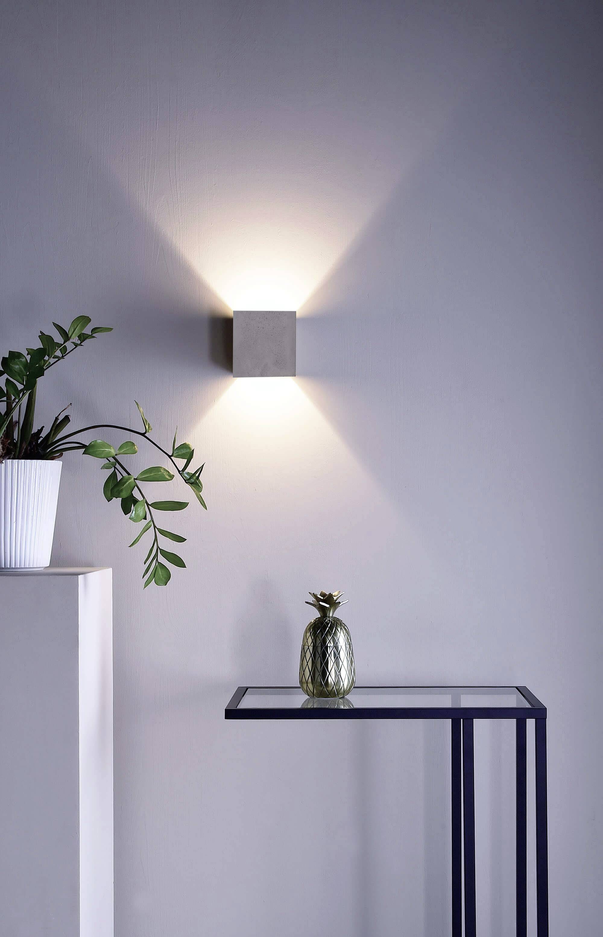 Nástenná lampa vyžaruje teplé svetlo. Vedľa nej je malý stolík so skleným povrchom, na ktorom stojí dekorácia v podobe ananásu. Vľavo stojí rastlina v bielom kvetináči.