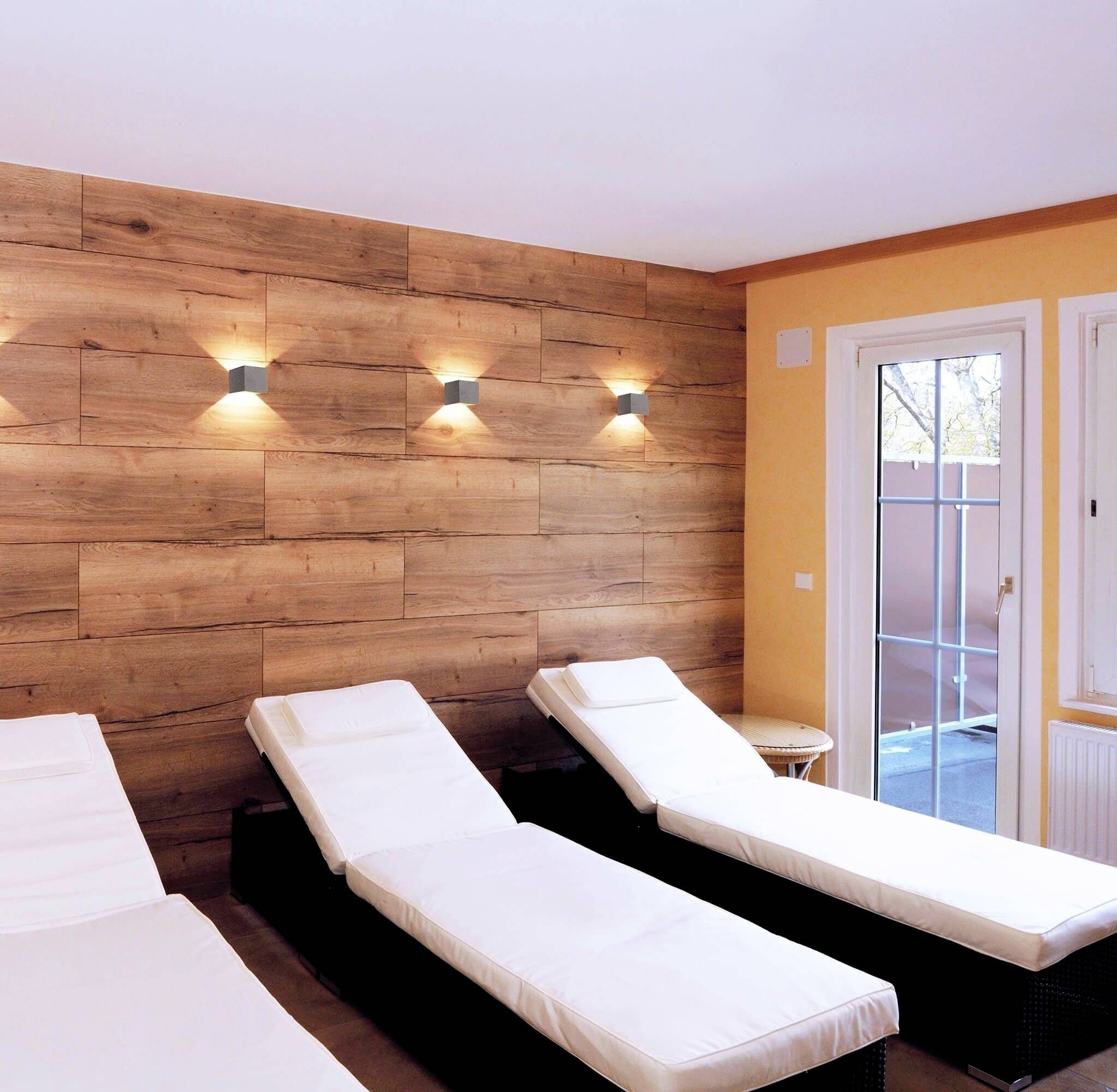 Relaxačná miestnosť so štyrmi bielymi ležadlami na hnedej podlahe, drevená stena s osvetlenými lampami, okno na pravej strane.