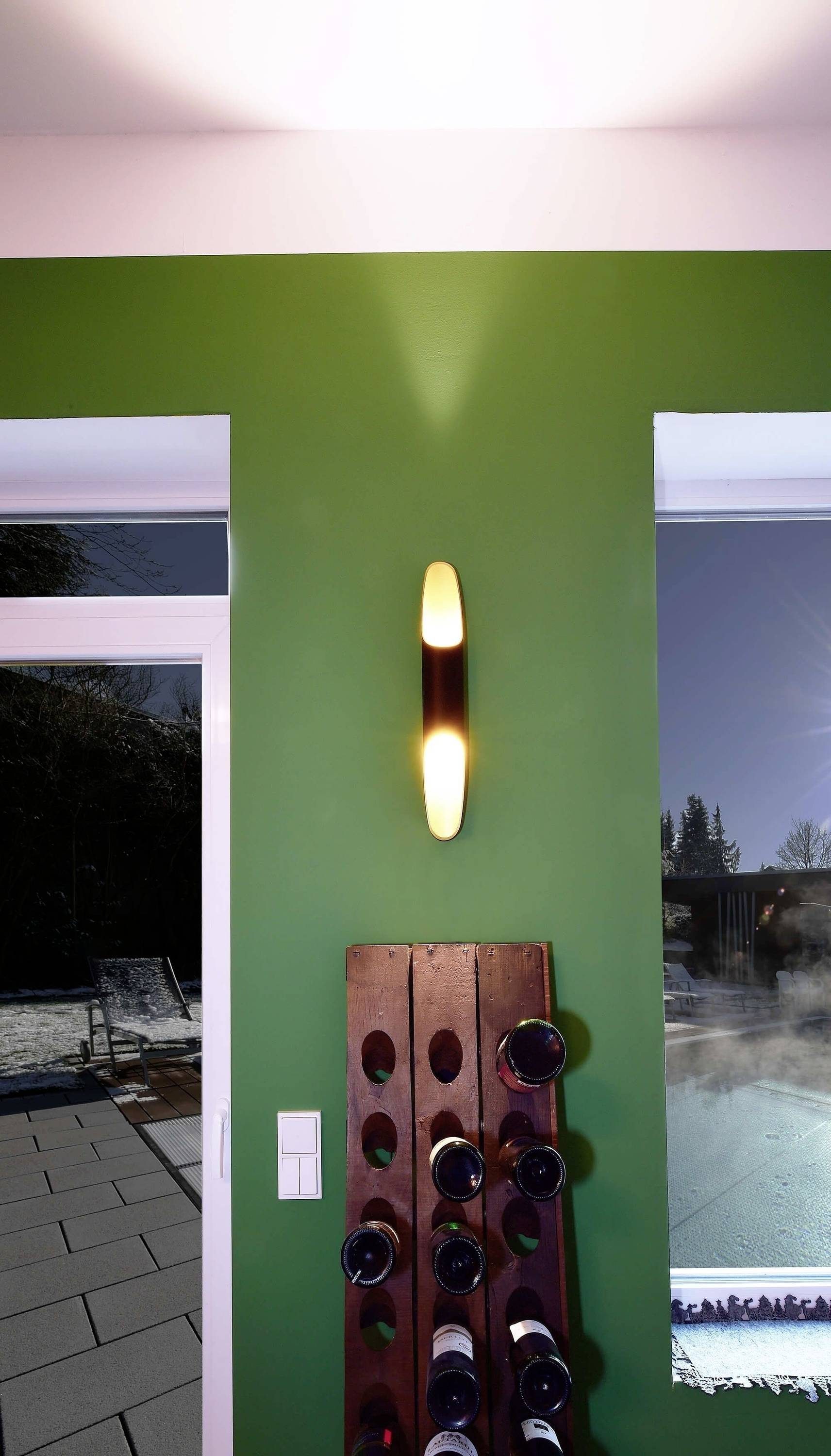 Nástenná lampa na zelenej stene nad regálom s vínom, s výhľadom na terasu a zimná záhrada.