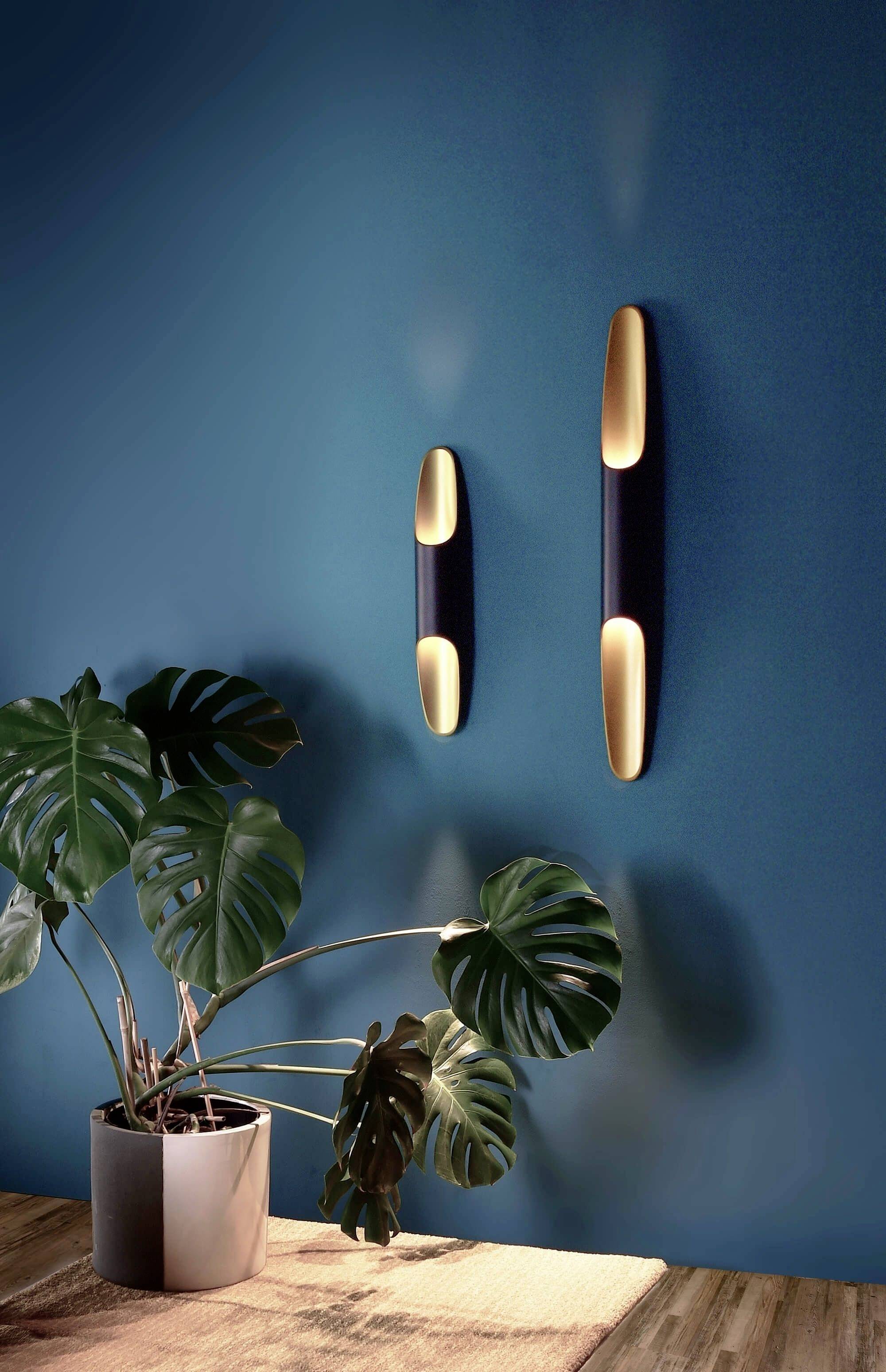 Izba s múrom, na ktorom sú zavesené dve moderné nástenné lampy. Pod nimi stojí rastlina v kvetináči na drevenom stolíku.