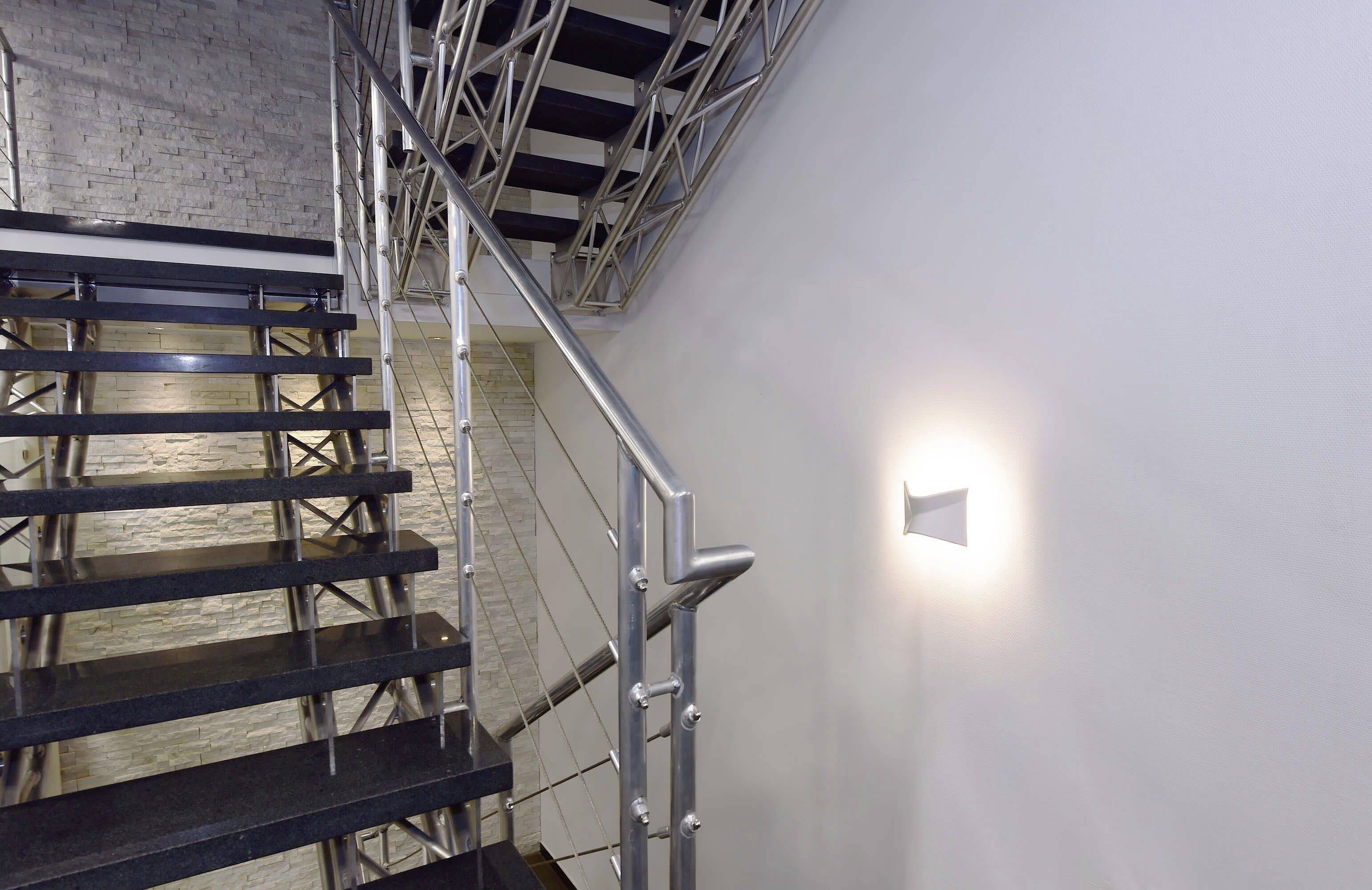 Moderné kovové schodisko s tmavými stupňami vedie nahor pozdĺž steny s žltým svetlom, ktoré vrháva geometrický tieň.