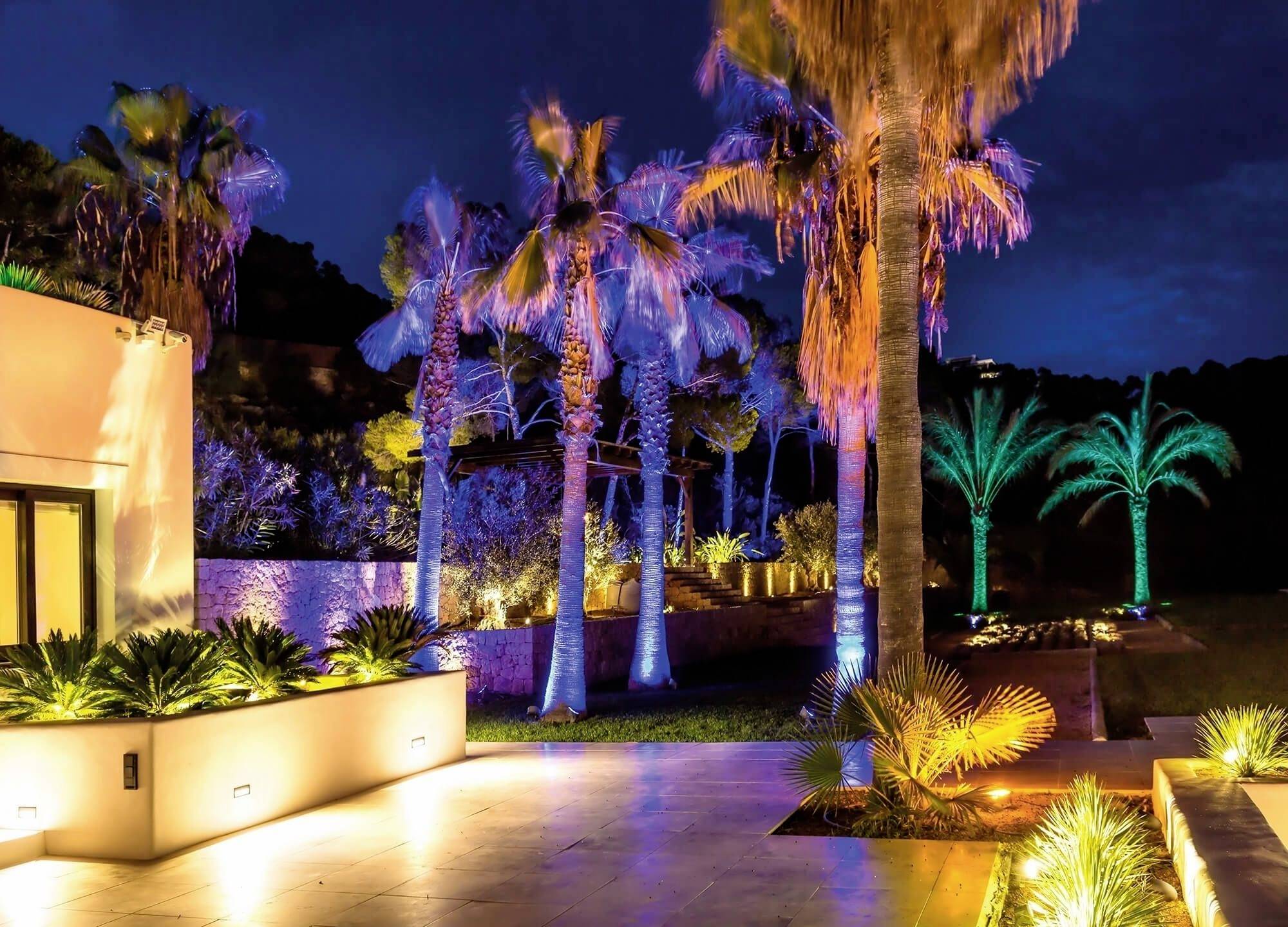 Večerná záhradná krajina s osvetlenými palmami a terasou. Dom vľavo v obraze. Tropická atmosféra v tmavom prostredí.
