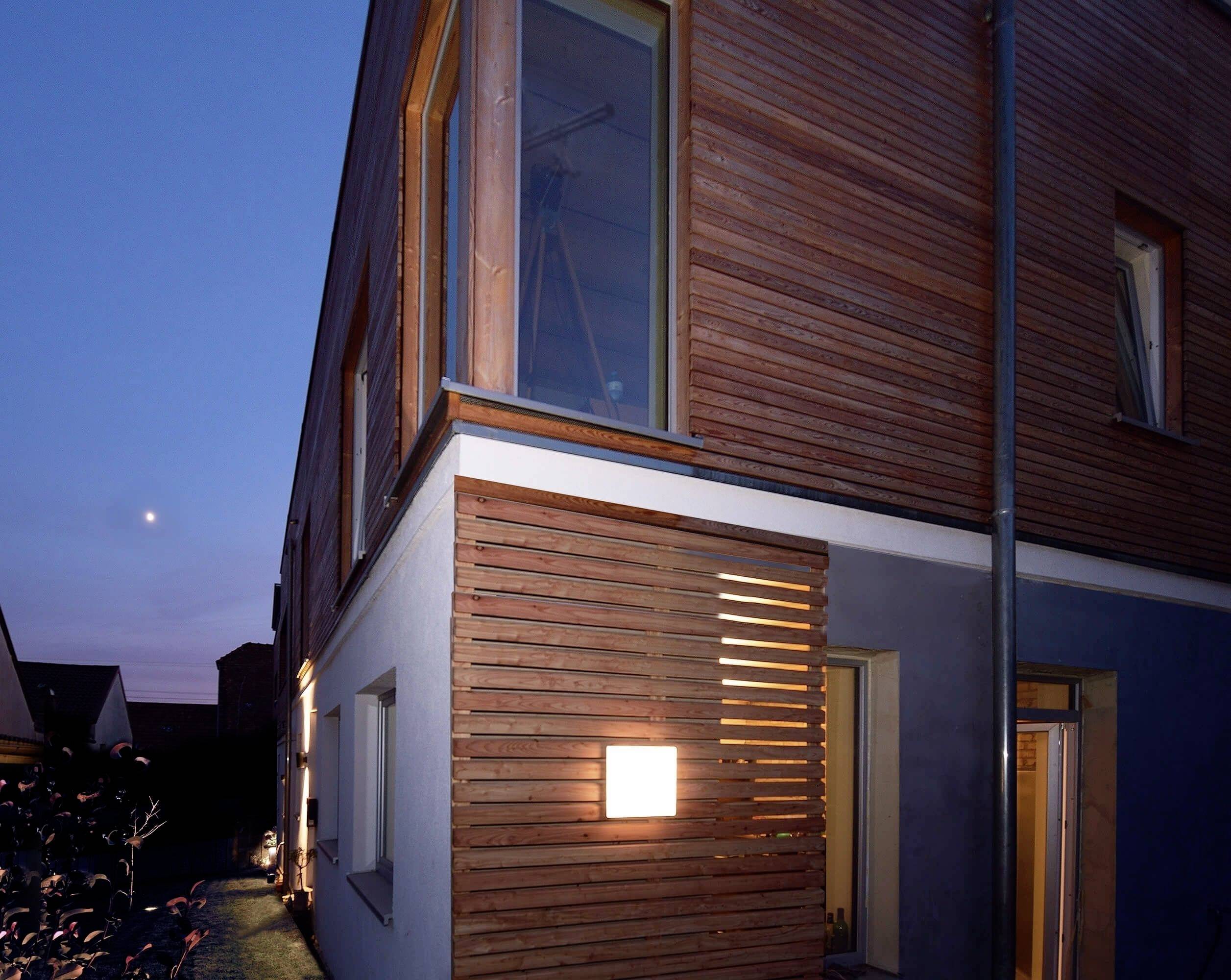 Pohľad z boku na moderný dom za súmraku, s dreveným obkladom a osvetleným oknom. Obloha je jemne tmavmodrá.