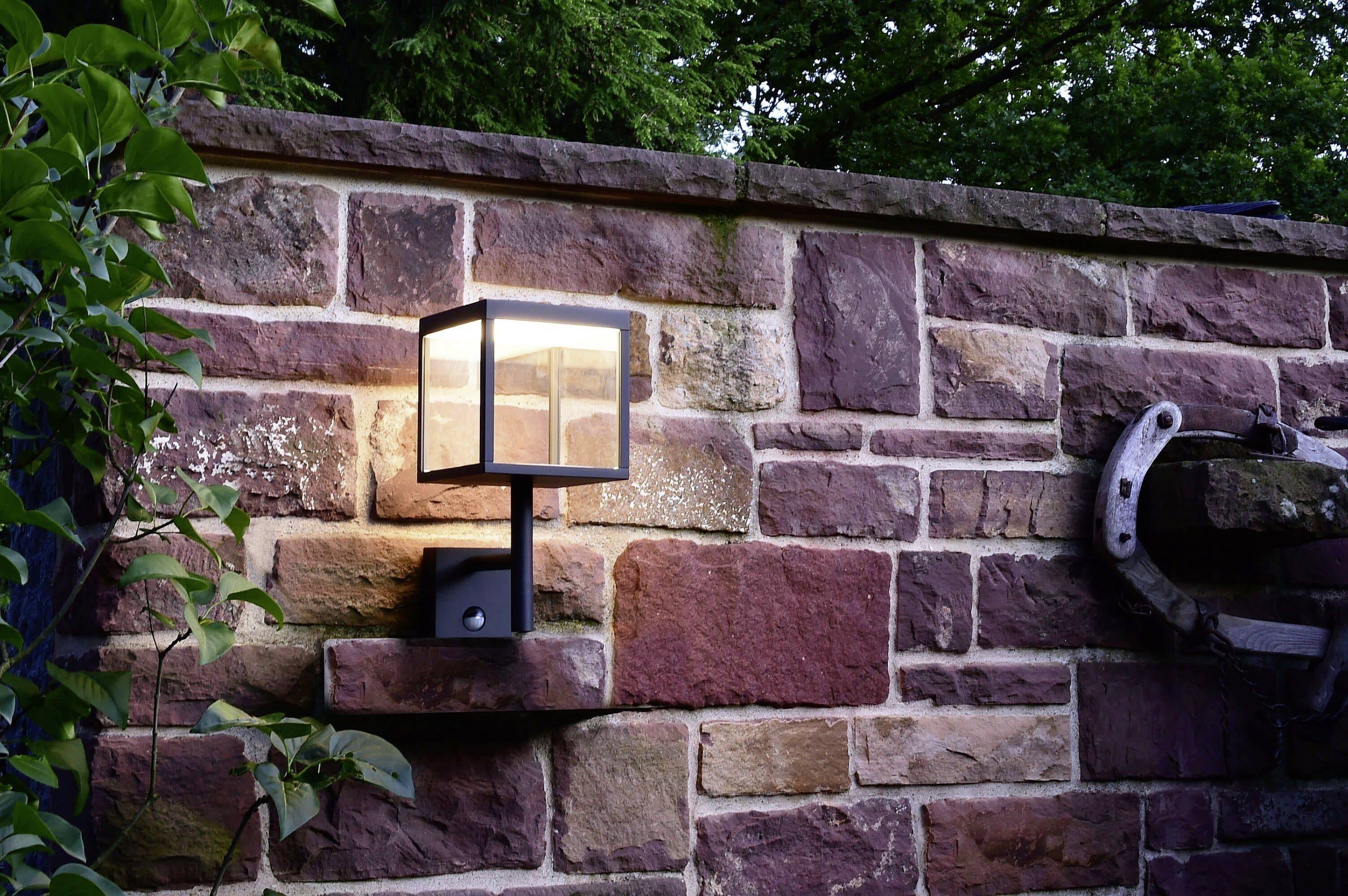 Nástenná lampa svieti na rustikálnej kamennej stene, obklopená rastlinami. Scéna vytvára útulnú atmosféru záhrady.