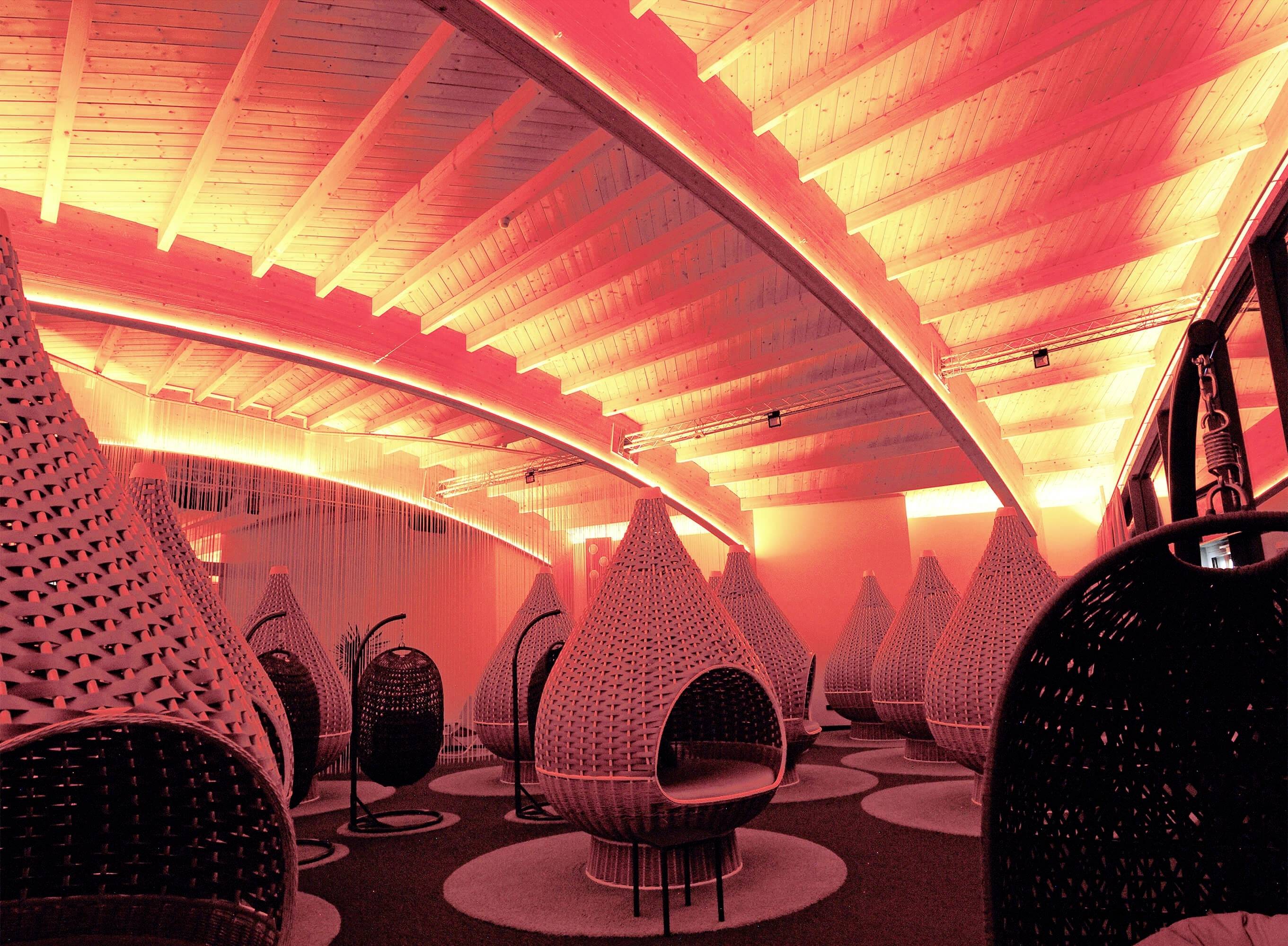 Štýlový priestor so červenými svetelnými efektmi, obsahujúci visiace, kvapkovité sedacie prvky z ľahkého a tmavého pletiva na okrúhlych kobercoch.