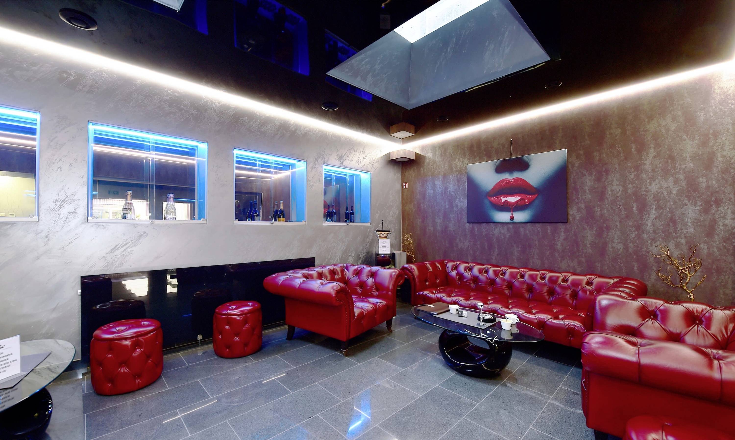 Elegantný priestor lounge s červenými koženými sedačkami a taburetkami, moderným umením na stene a modrým osvetlením.