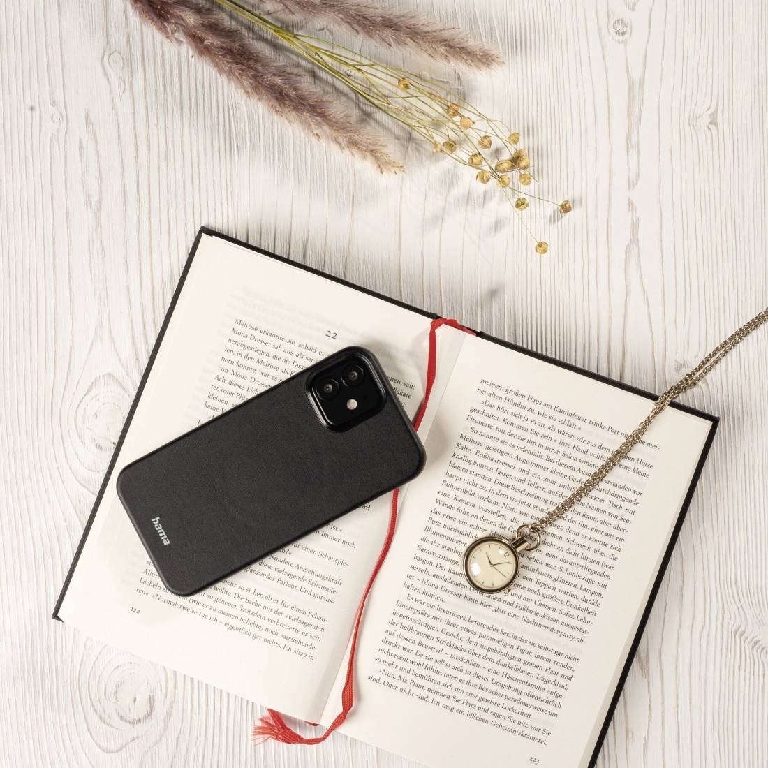 Otvorená kniha na drevenej doske, na ktorej sú umiestnené čierne smartfón a vreckové hodinky, dekorované sušenými rastlinami.